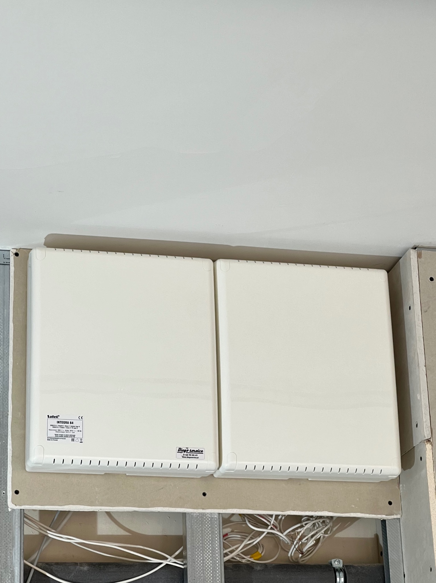 Dwa białe panele centrali alarmowej Satel Integra 64 zamontowane na płycie gipsowo-kartonowej, widoczne kable instalacji elektrycznej.