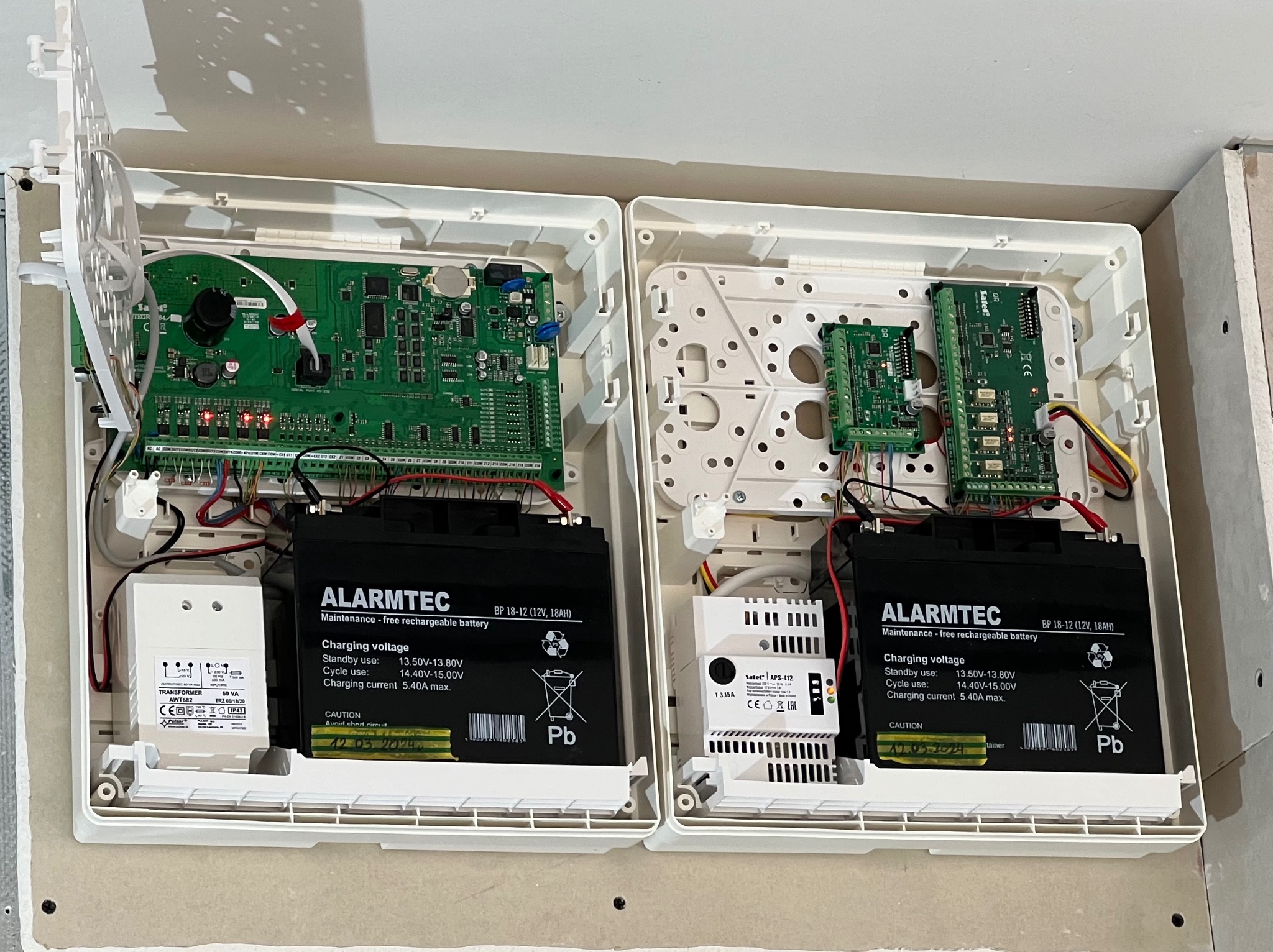 Otwarta obudowa centrali alarmowej z widocznymi modułami elektronicznymi, akumulatorami podtrzymującymi i transformatorem zasilającym.