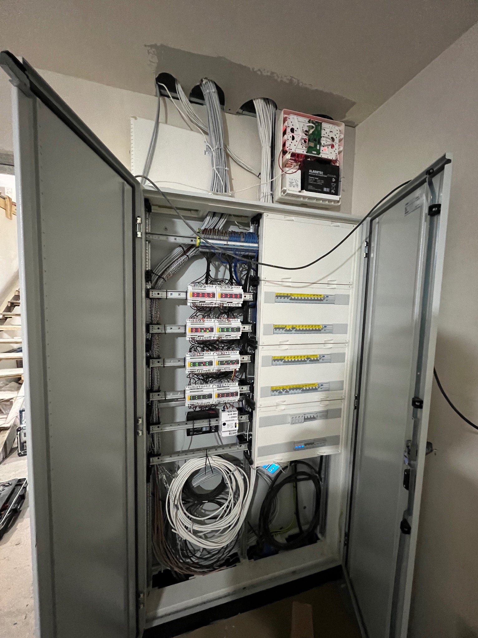 Otwarta szafa elektryczna z widocznymi bezpiecznikami, kablami i innym osprzętem, zamontowana w budynku w Przyprostyni, z widocznymi śladami prac wykończeniowych.