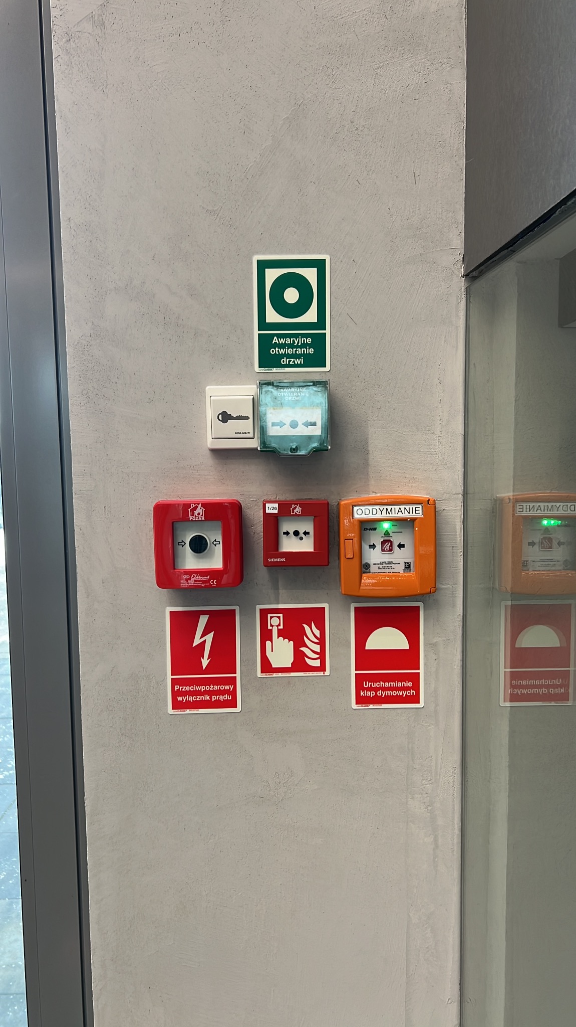 Zestaw różnych przycisków i znaków bezpieczeństwa na ścianie, w tym przycisk awaryjnego otwierania drzwi, wyłącznik przeciwpożarowy prądu, przycisk oddymiania, uruchamianie klap dymowych...