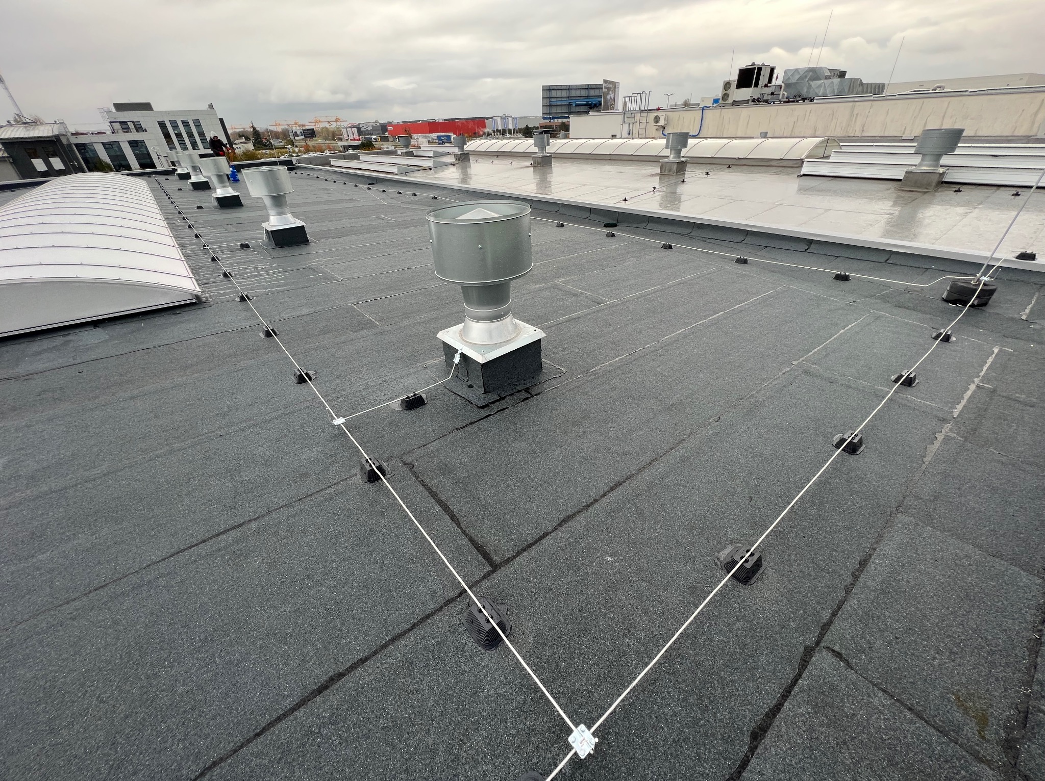 Płaski dach pokryty papą z zainstalowaną instalacją odgromową, widoczne metalowe kominki wentylacyjne i przewody odgromowe z uchwytami, w tle budynki przemysłowe i pochmurne niebo.