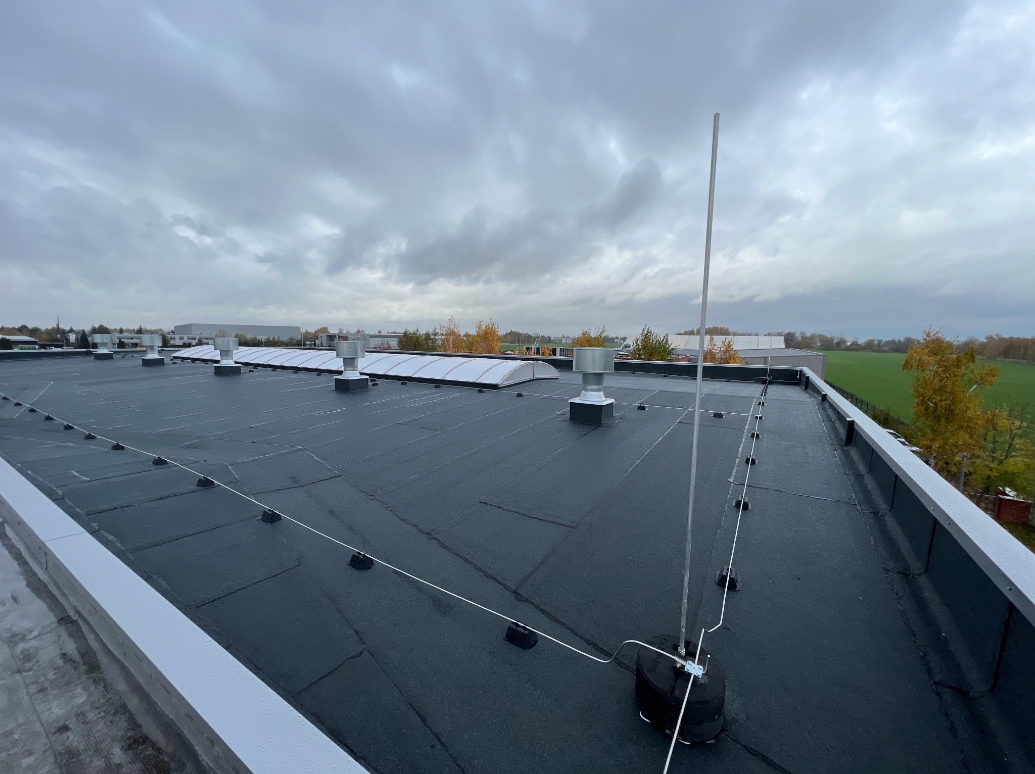 Widok z góry na płaski dach budynku przemysłowego z zainstalowaną instalacją odgromową, widoczne przewody i maszt odgromowy na tle pochmurnego nieba.