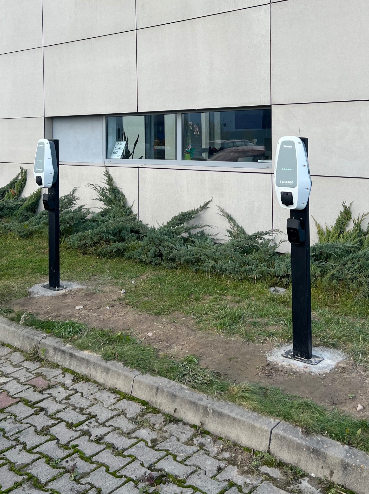 Dwa stanowiska ładowania samochodów elektrycznych, model I-CHARGE, zamontowane przed budynkiem z szarą elewacją, widoczne krzewy i chodnik z kostki brukowej.
