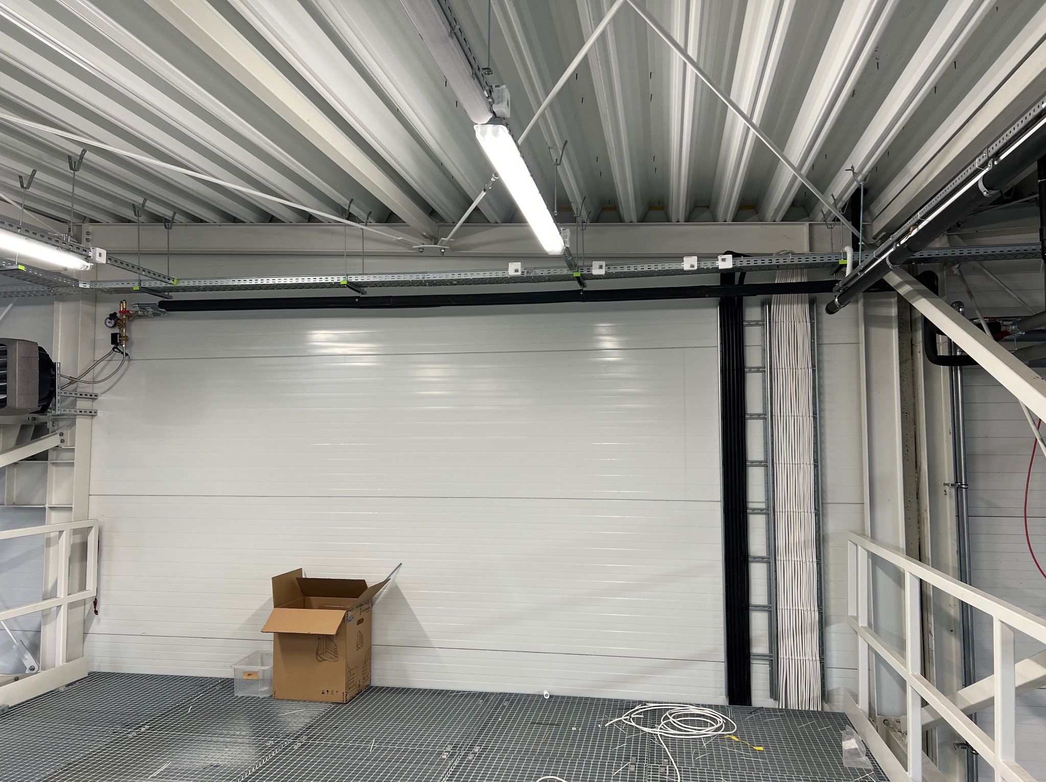 Wnętrze hali produkcyjnej z widoczną białą bramą segmentową, instalacją elektryczną w korytach kablowych pod sufitem, oraz rozłożonymi kablami na metalowej podłodze kratownicowej. W tle widoczny...