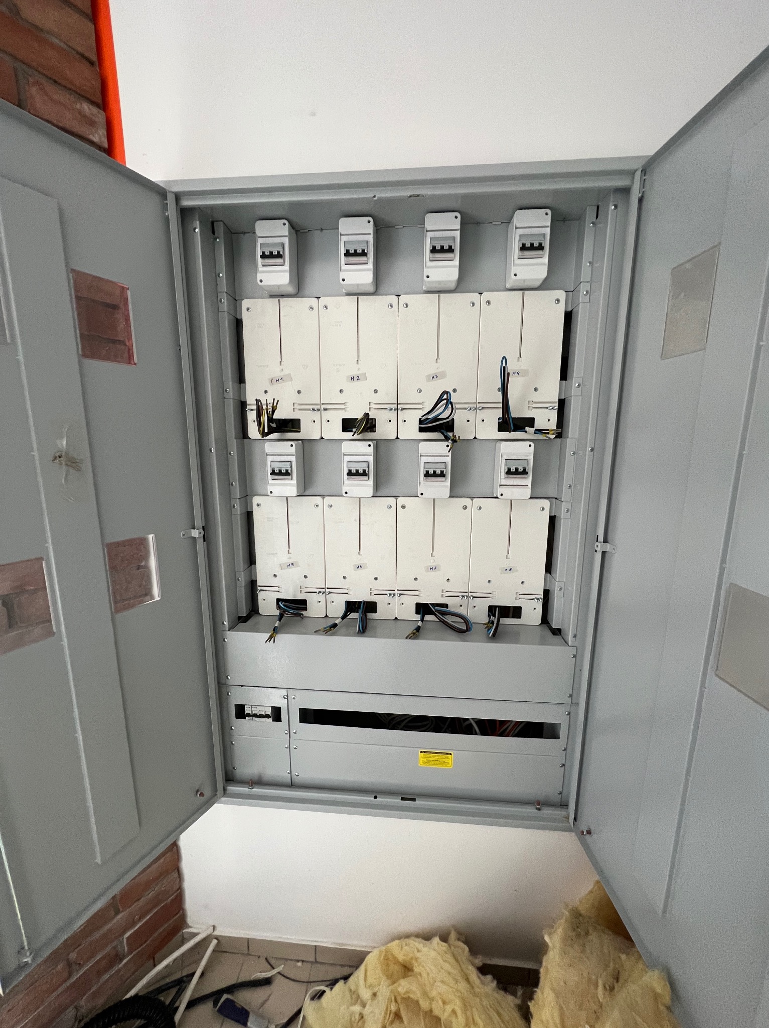 Otwarta skrzynka elektryczna z zainstalowanymi bezpiecznikami i podłączonymi przewodami, widoczne fragmenty muru z cegły i izolacji.