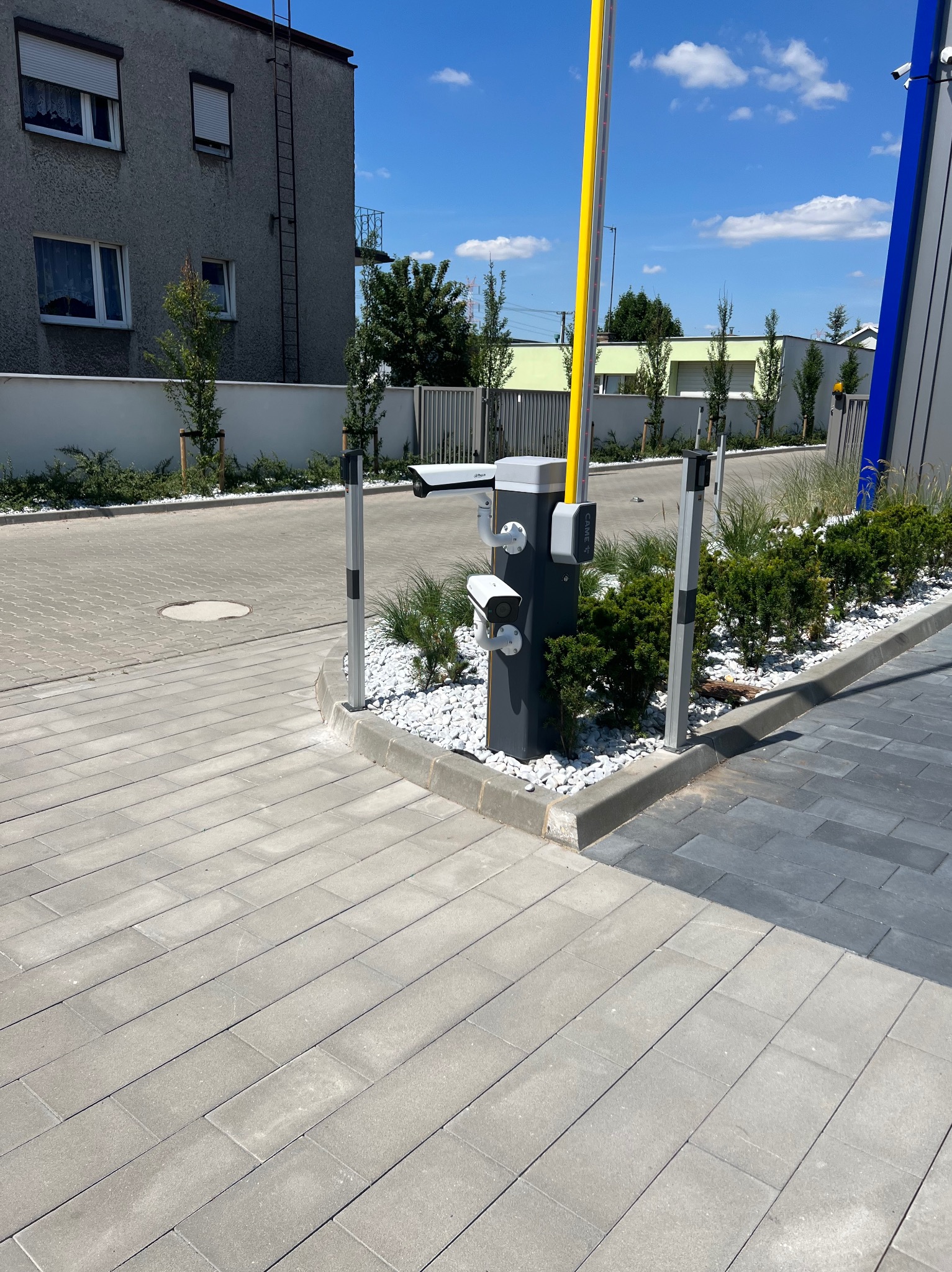 Automatyczna szlaban z żółtą belką w pozycji pionowej, kamery monitoringu na słupku, otoczenie wyłożone kostką brukową i ozdobione białymi kamieniami oraz zielenią.