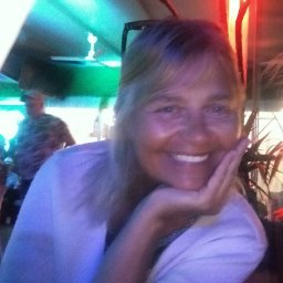 Trzeźwy Azyl Anna Rewekant - Uśmiechnięta kobieta z ręką pod brodą, w tle widoczny mężczyzna i elementy wystroju wnętrza, oświetlone kolorowymi światłami.