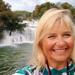 Trzeźwy Azyl Anna Rewekant - Uśmiechnięta blond kobieta na tle wodospadu w Parku Narodowym Krka, Chorwacja. Kadr z bliska, widoczny fragment drewnianej barierki.