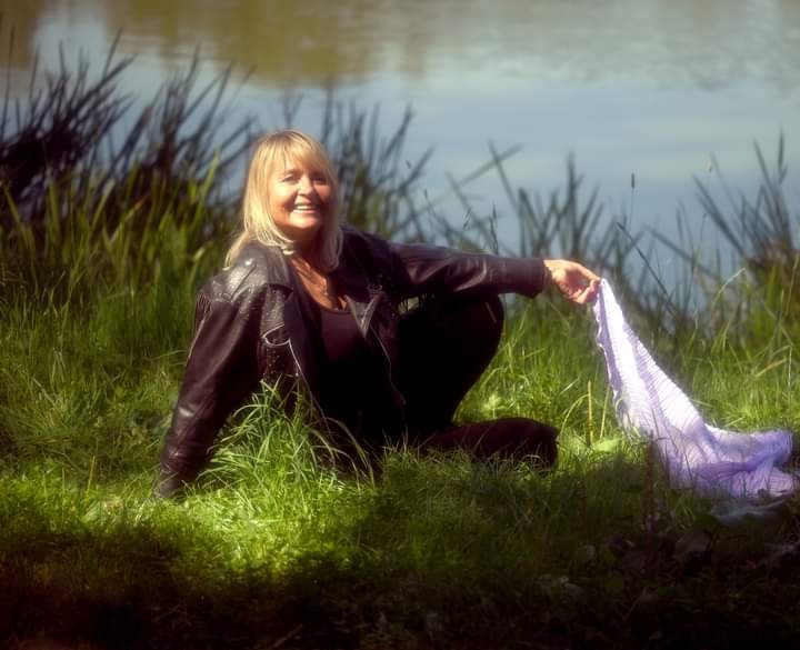 Uśmiechnięta blond kobieta w skórzanej kurtce siedzi w trawie nad brzegiem wody, trzymając w dłoni jasnofioletową chustę.