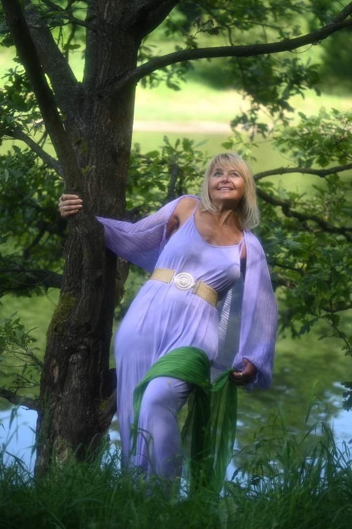 Uśmiechnięta blond kobieta w liliowej sukience, opierająca się o drzewo w lesie, z zielonym szalem w ręku i widocznym jeziorem w tle.