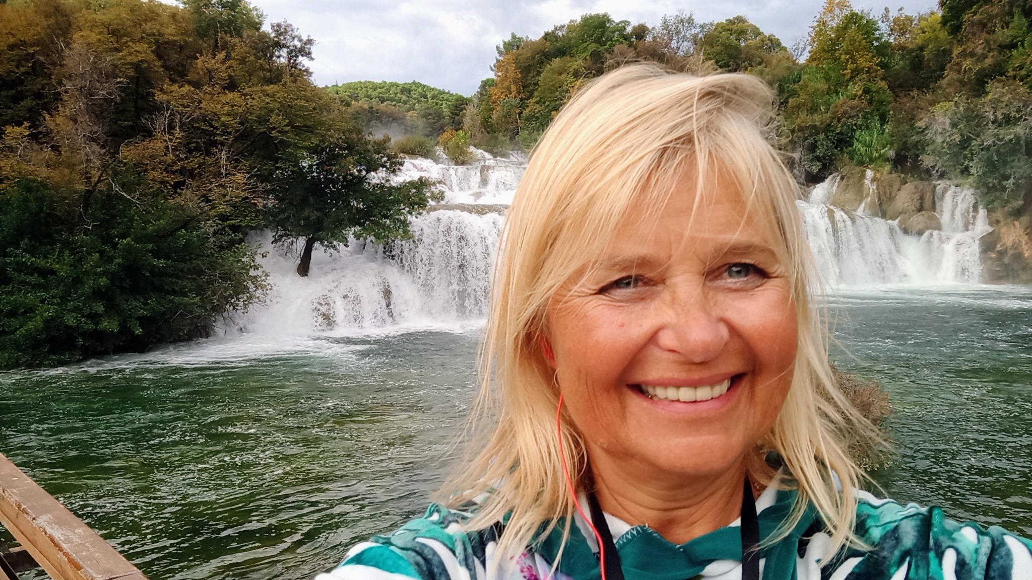 Uśmiechnięta blond kobieta na tle wodospadu w Parku Narodowym Krka, Chorwacja. Kadr z bliska, widoczny fragment drewnianej barierki.