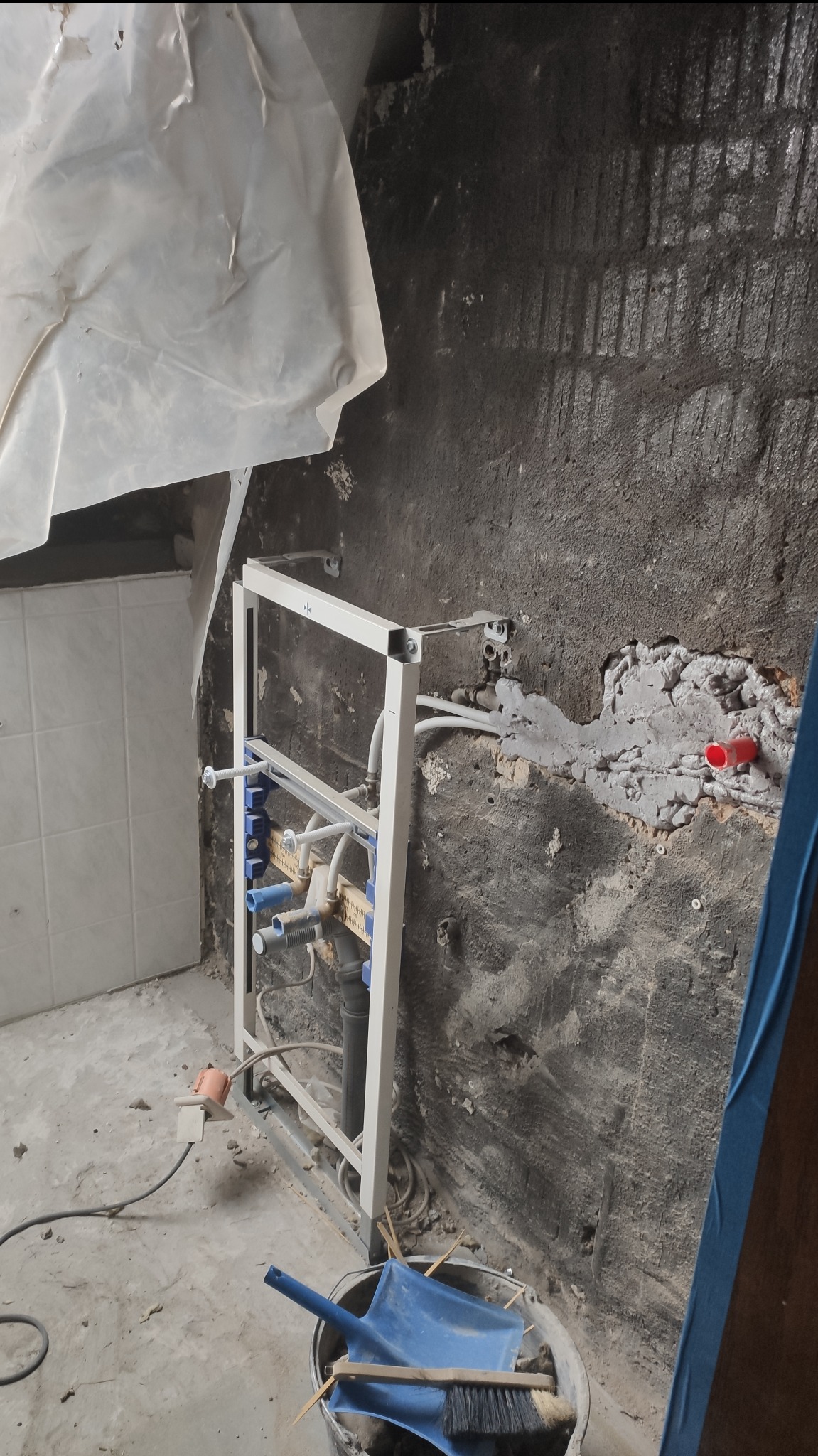 Stelaż podtynkowy do WC zamontowany na surowej ścianie w trakcie remontu łazienki, widoczne instalacje wodne i elektryczne, wiadro z narzędziami w tle.