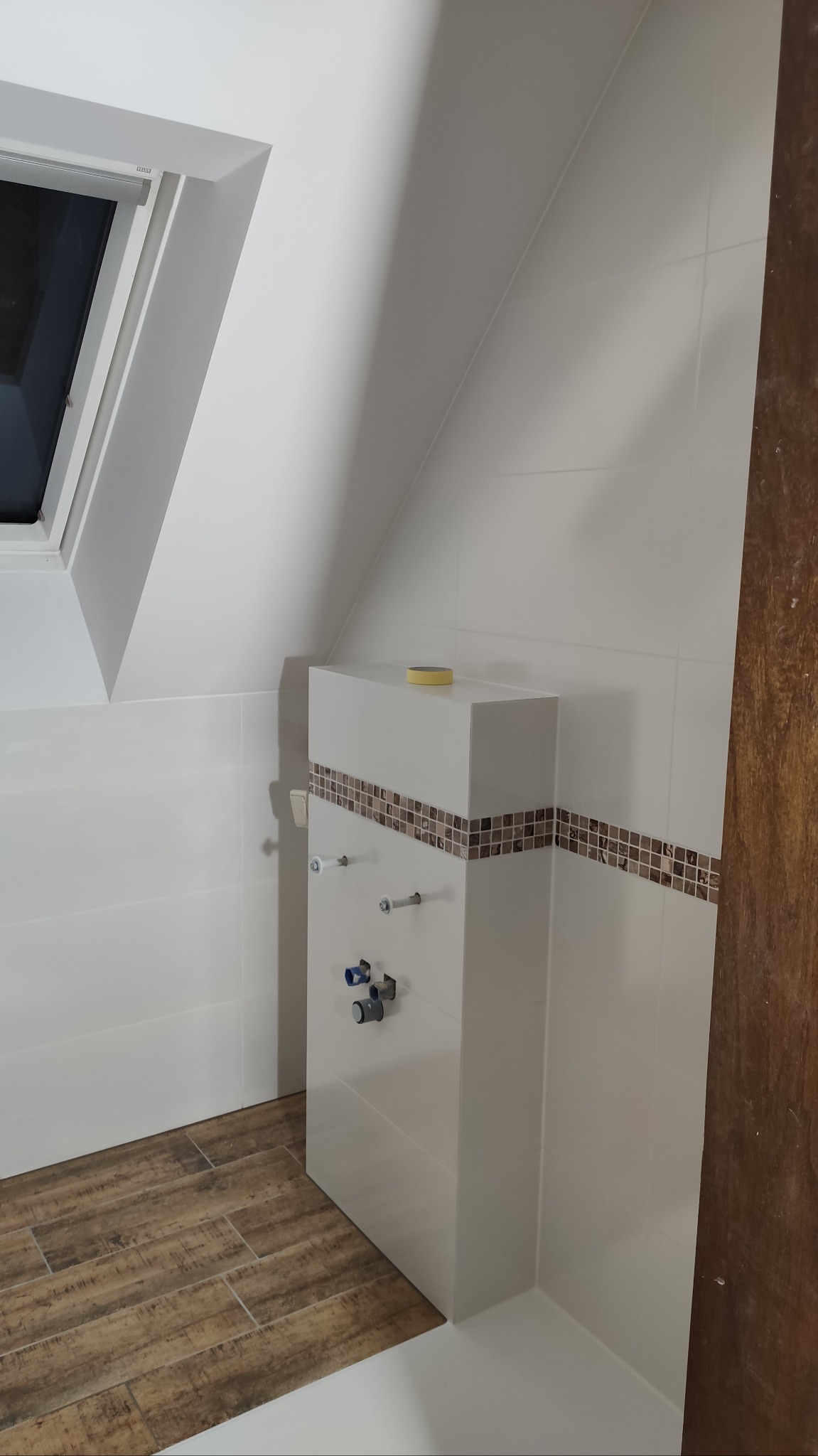 Biała zabudowa WC z mozaiką w łazience na poddaszu, widoczne przyłącza wodne i okno dachowe.