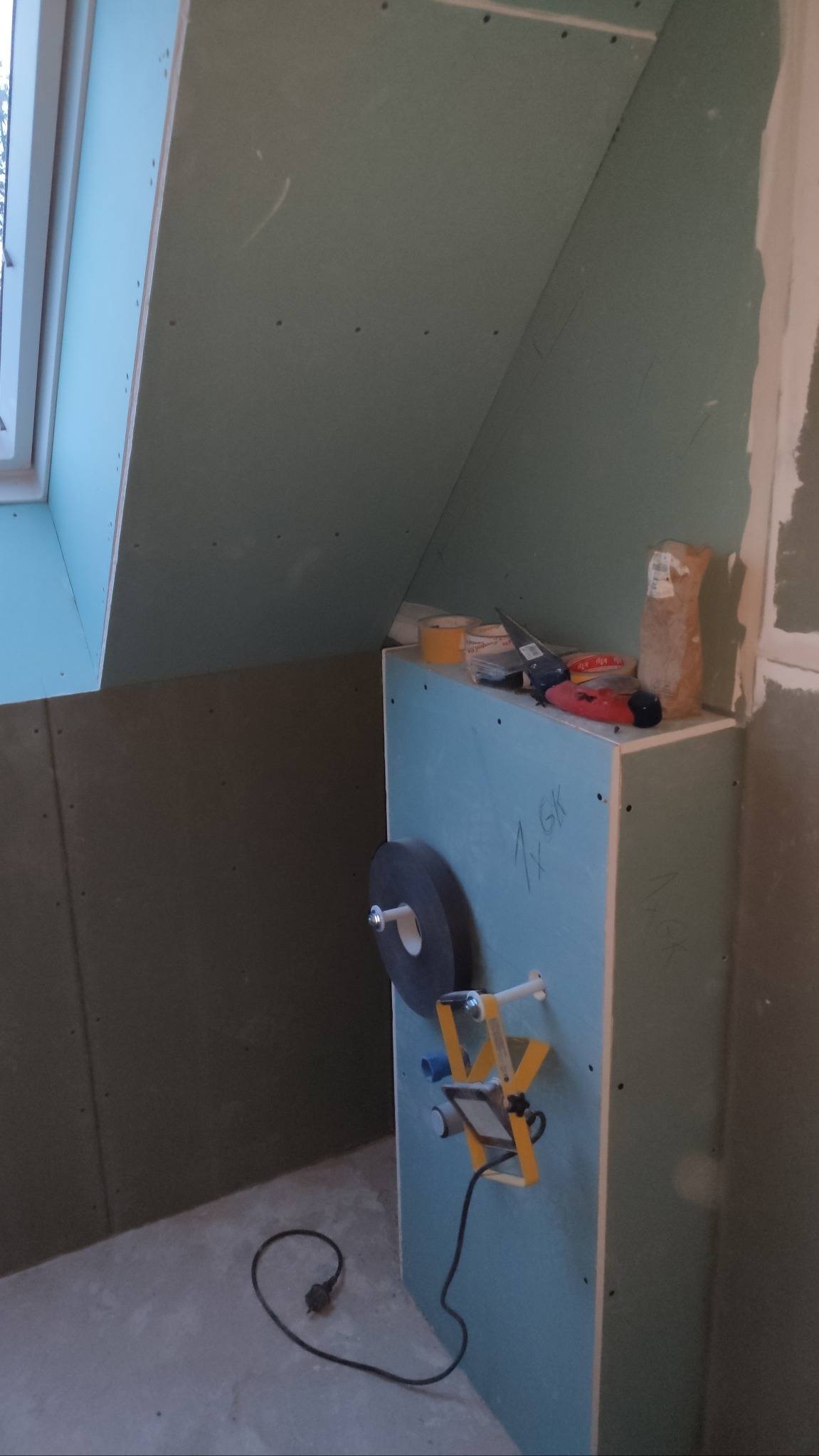 Montaż płyt gipsowo-kartonowych na poddaszu, widoczna konstrukcja z profili, szpachla, taśma, narzędzia i lampa budowlana na prowizorycznym postumencie z płyty GK.