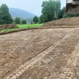 Przygotowany teren pod budowę z widocznymi śladami opon ciężkiego sprzętu, w tle zalesione wzgórza i fragment łyżki koparki w prawym górnym rogu.