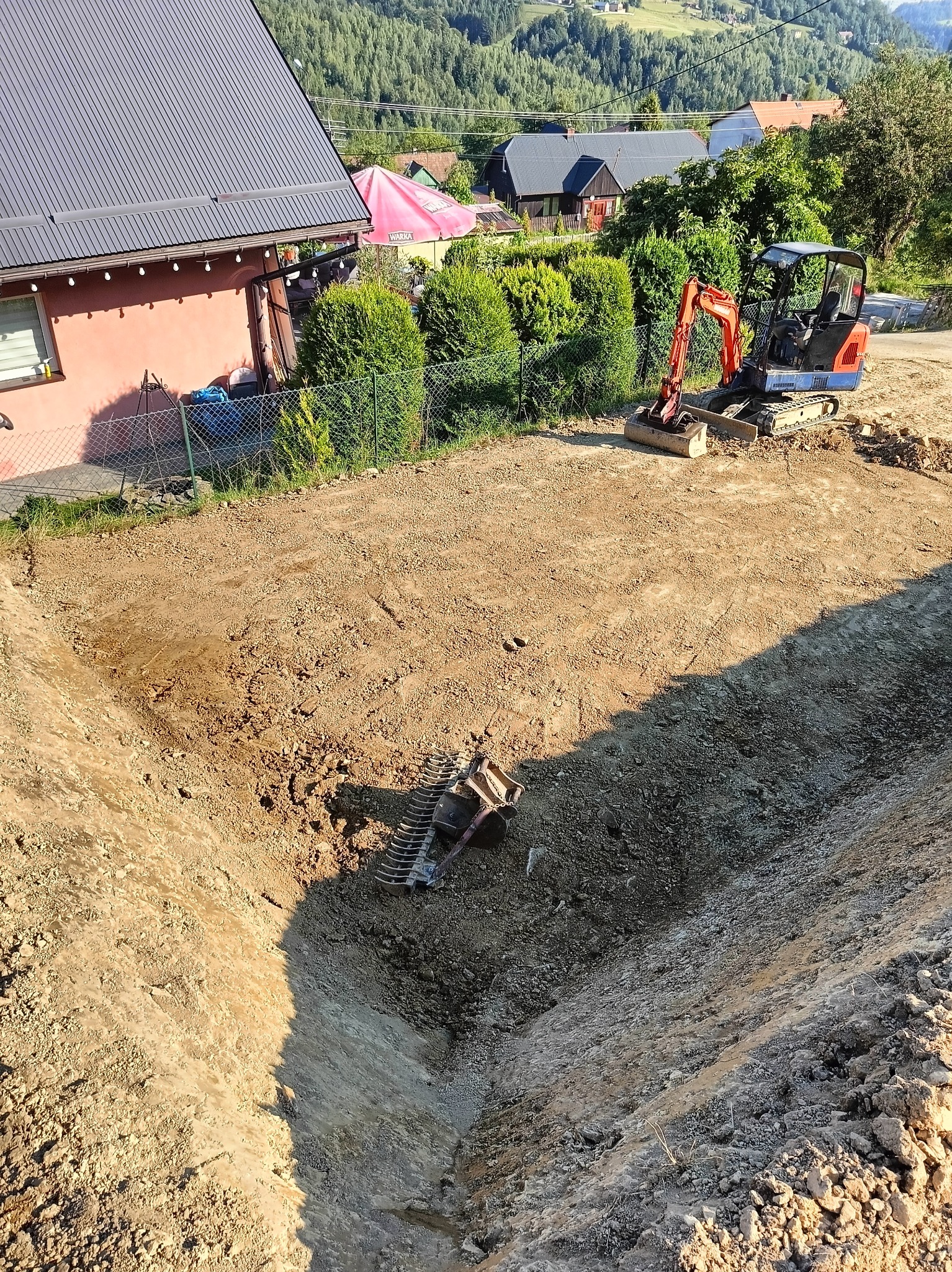 Głęboki wykop w ziemi z widoczną łyżką koparki na dnie, mała koparka stoi obok wykopu na tle zabudowań i zieleni.