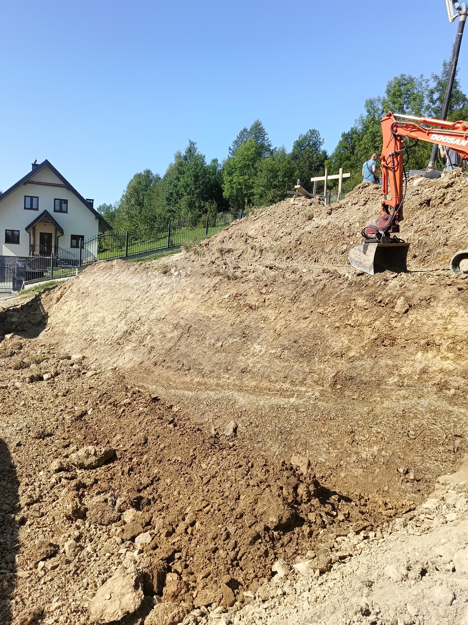 Pomarańczowa koparka Doosan na zboczu ziemi, w tle dom jednorodzinny i pracownik przy wyznaczaniu terenu.