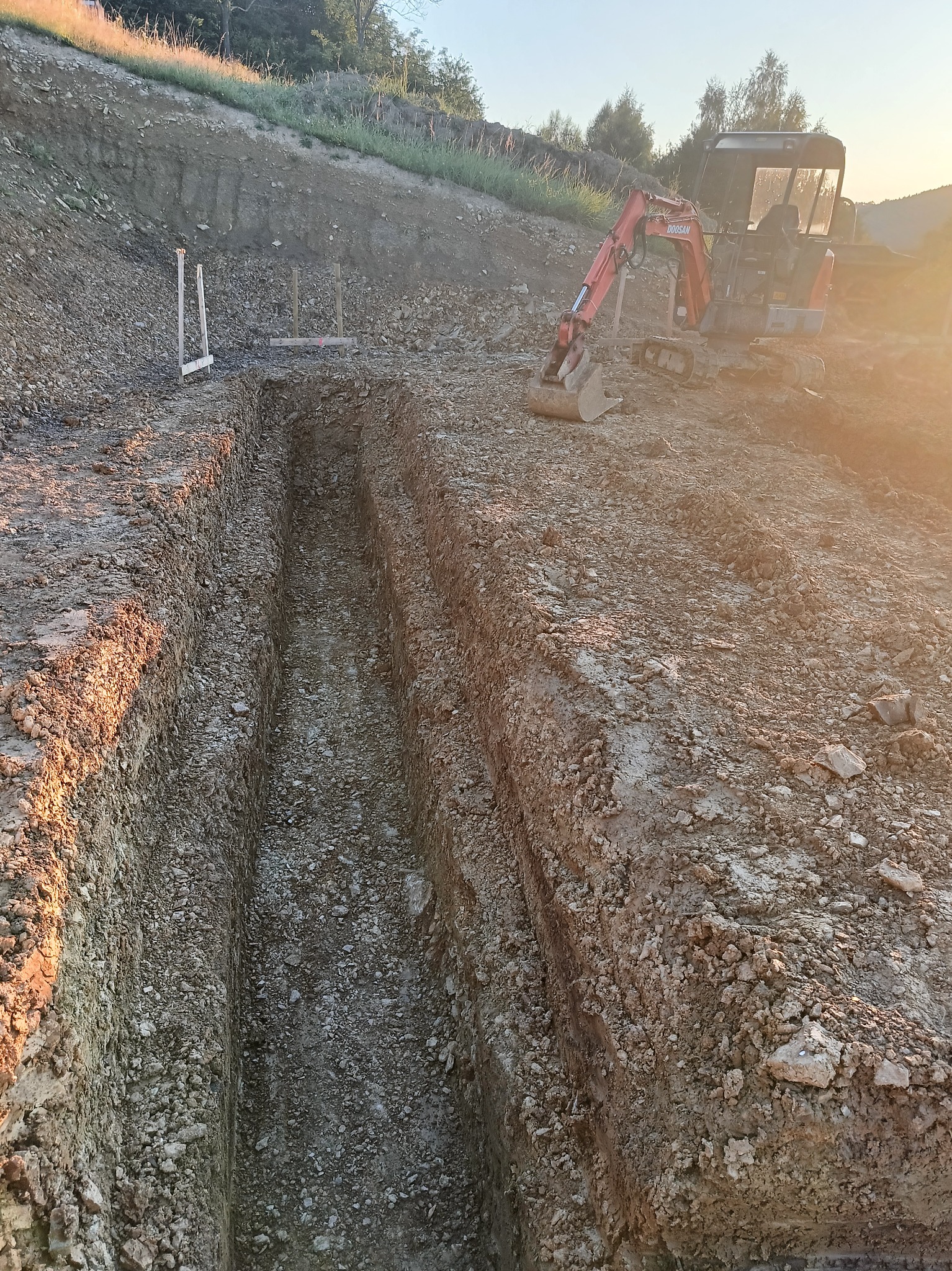 Wąski wykop pod instalacje, widoczny z góry, z mini koparką Doosan w tle i drewnianymi znacznikami po lewej stronie