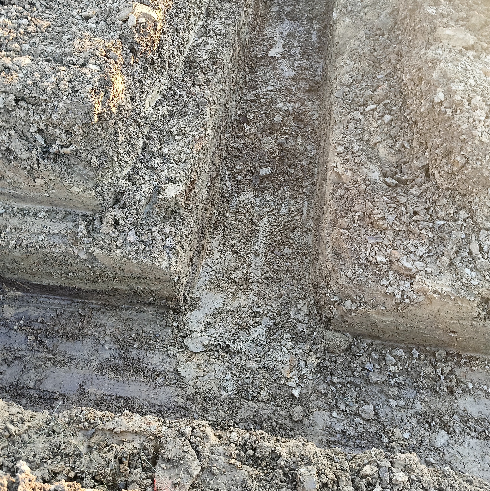 Wąski wykop w ziemi z widoczną strukturą gleby, kamieniami i śladami wilgoci na dnie.