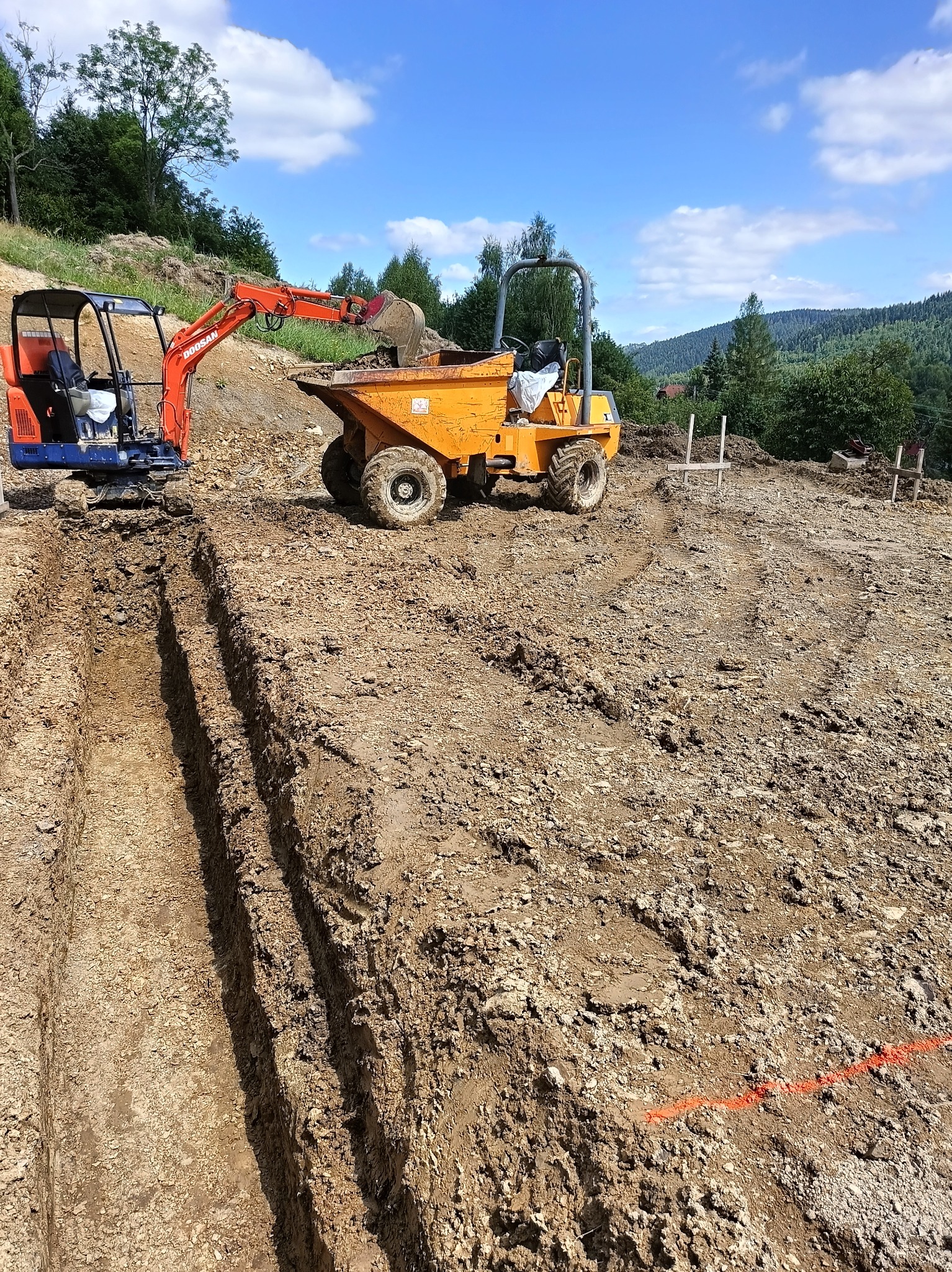 Pomarańczowy minikoparka Doosan nabiera ziemię do wywrotki na placu budowy, widoczne wykopane rowy i tyczki geodezyjne, w tle zalesione wzgórza pod błękitnym niebem.