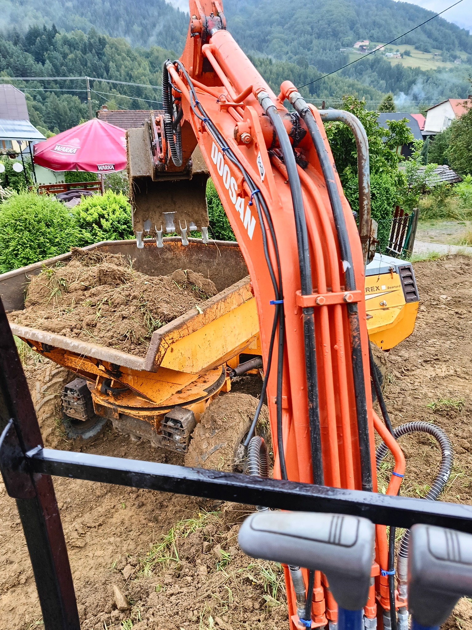 Pomarańczowa koparka Doosan na nierównym terenie, nabiera ziemię z trawą do żółtej wywrotki Rex Benford, widok z kabiny operatora, w tle zabudowania i zalesione wzgórza.