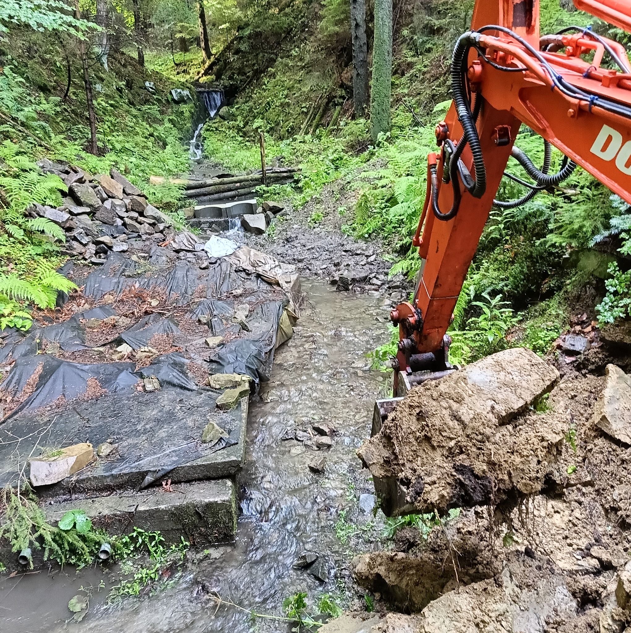Pomarańczowa koparka z łyżką pełną ziemi na brzegu potoku w lesie, betonowe elementy umocnienia brzegów, w tle mały wodospad i bujna roślinność.