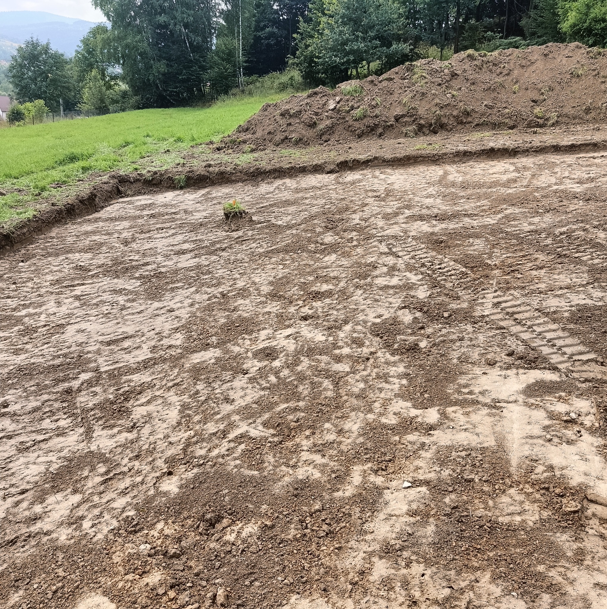 Przygotowany teren pod budowę, widoczne ślady opon ciężkiego sprzętu, z niewielkim kopcem ziemi i pozostałością trawy w rogu placu.