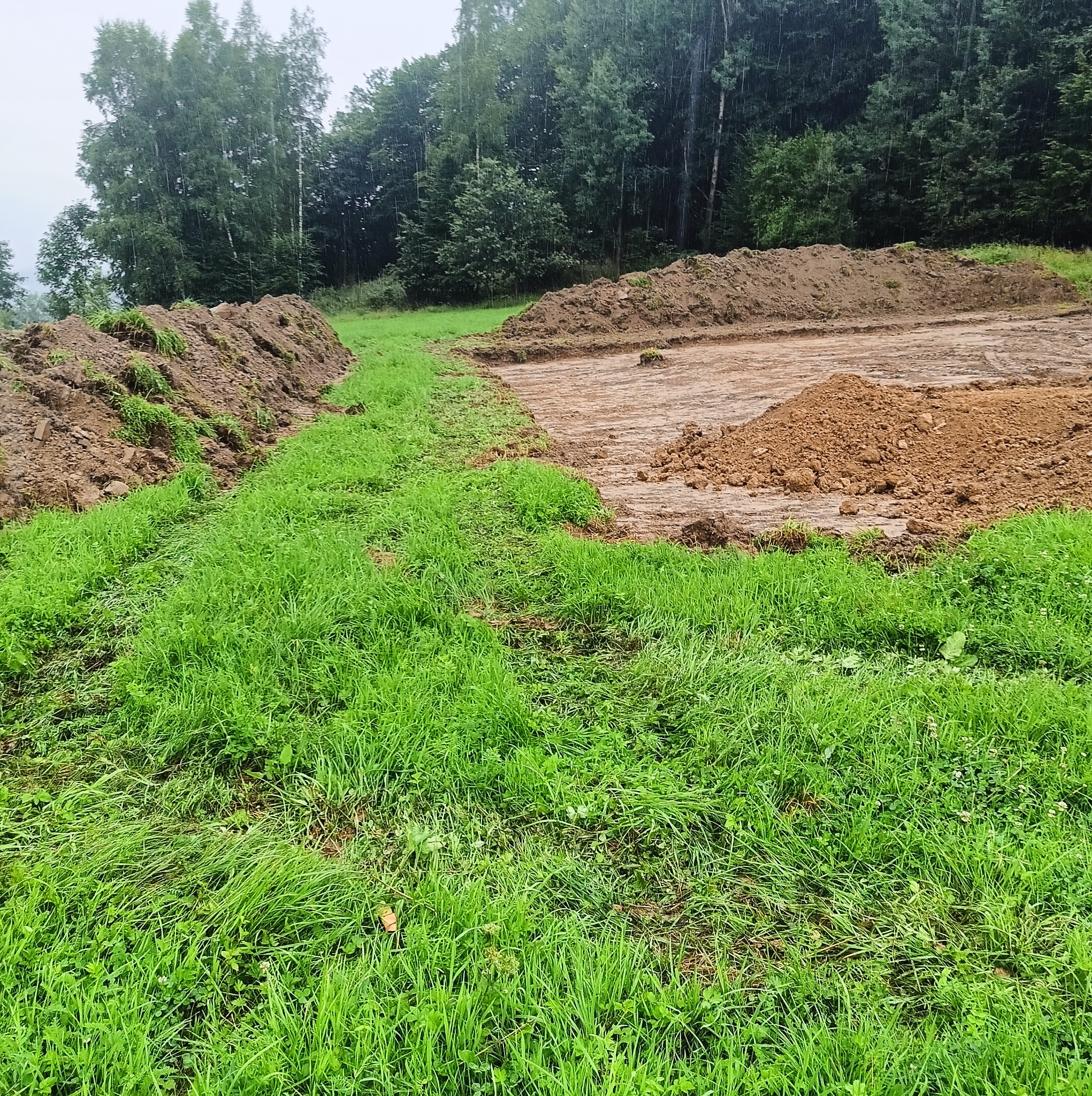 Przygotowany teren pod budowę z wykopem i usypanym brzegiem ziemi, widok na łąkę i las w tle.