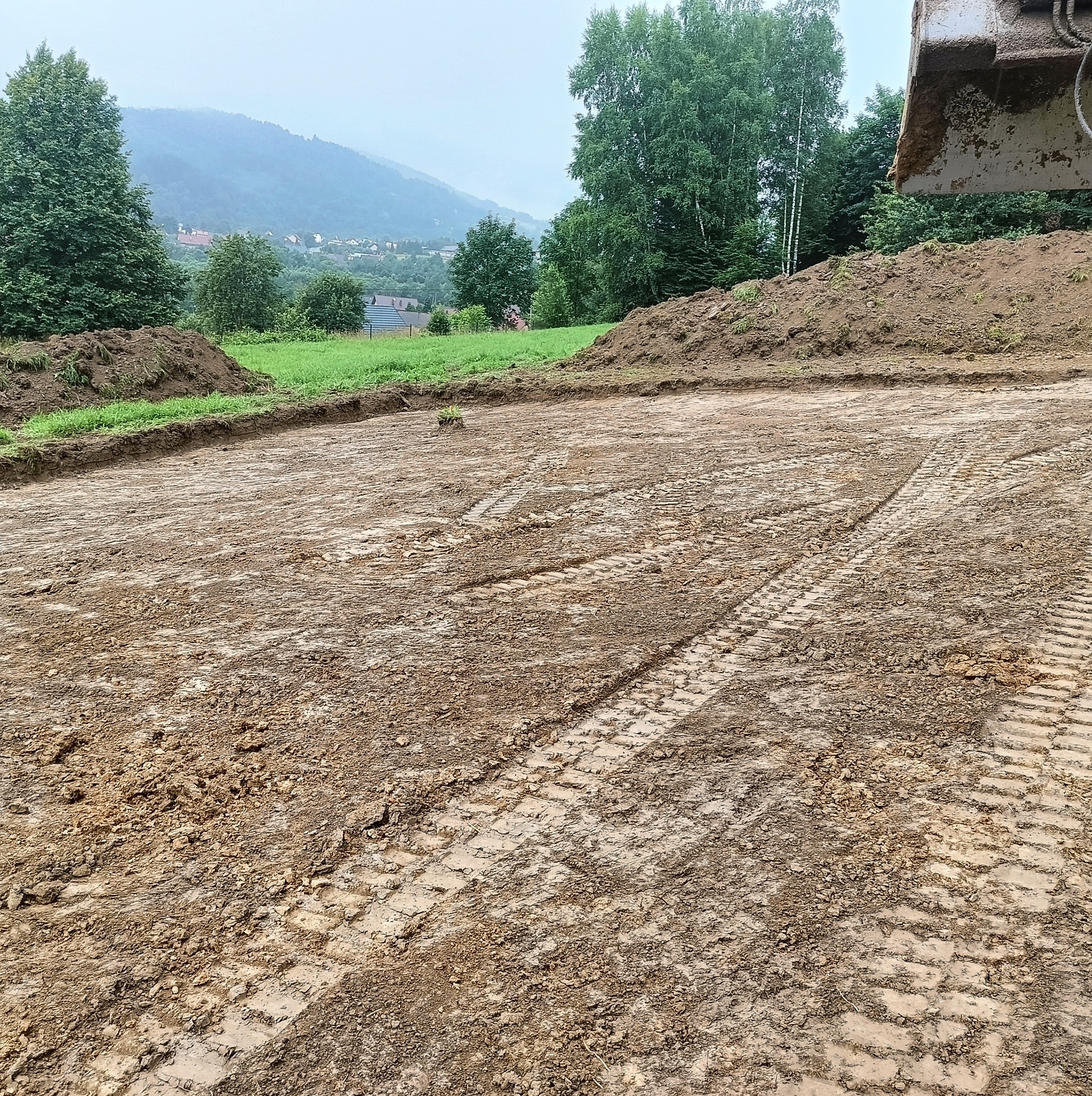 Przygotowany teren pod budowę z widocznymi śladami opon ciężkiego sprzętu, w tle zalesione wzgórza i fragment łyżki koparki w prawym górnym rogu.