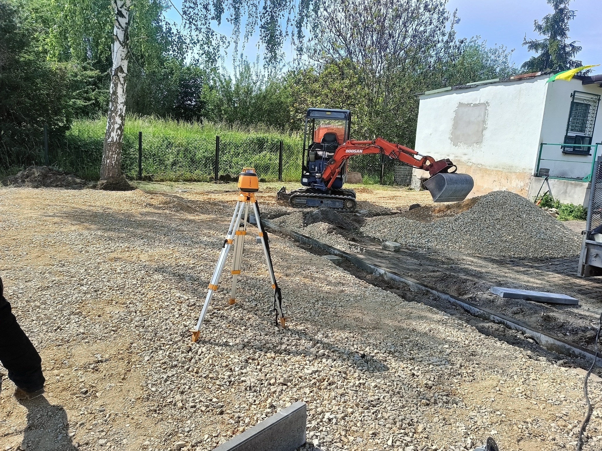 Mała koparka Doosan z łyżką, obok niwelator laserowy na statywie, świeżo wykopany rów z ułożonymi obrzeżami, nasypany żwir, w tle budynek i drzewa.