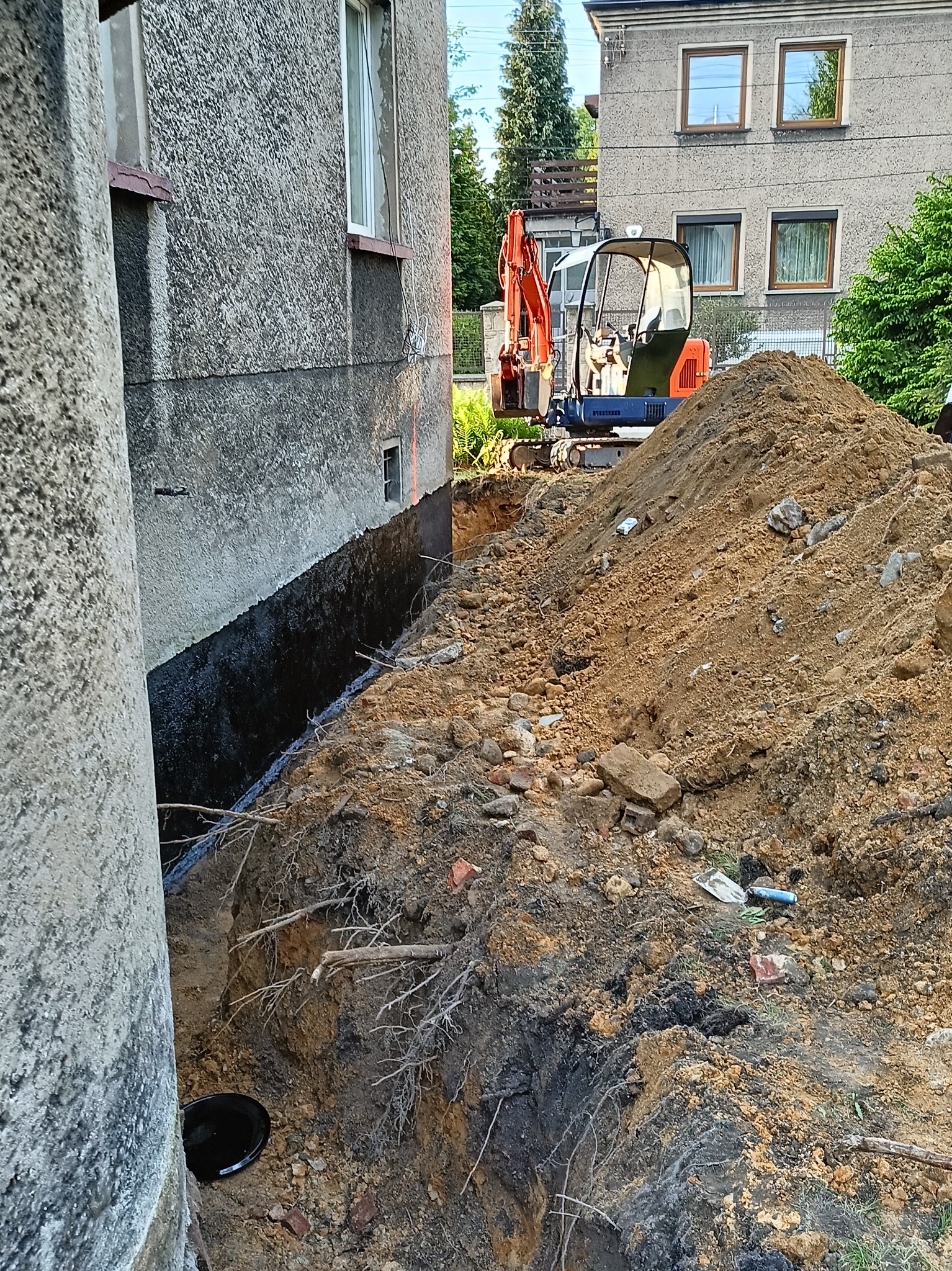 Wykop wzdłuż ściany budynku z widoczną warstwą izolacji, w tle pomarańczowa mini koparka i sterta ziemi.