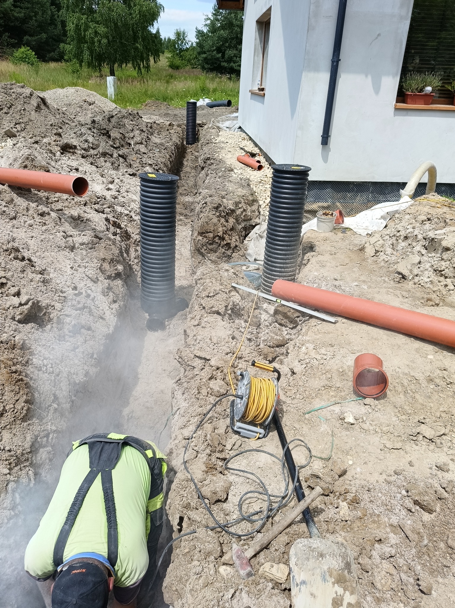 Pracownik w wykopie pod drenaż, widoczne rury drenarskie, narzędzia i fragment budynku w tle.