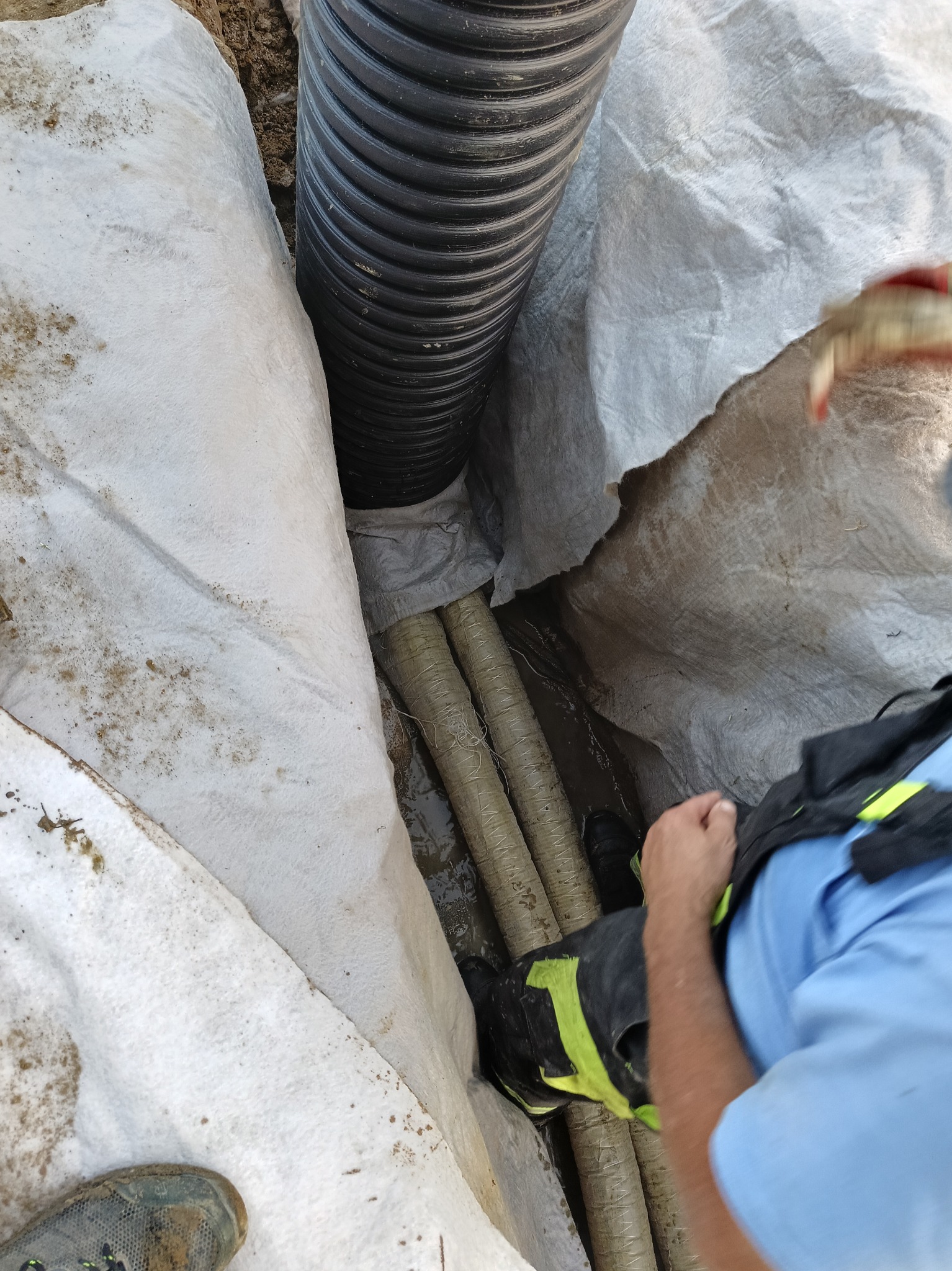 Czarny karbowany drenaż w wykopie wyłożonym białą geowłókniną, widoczne rury kanalizacyjne i fragment roboczego ubrania osoby nadzorującej.