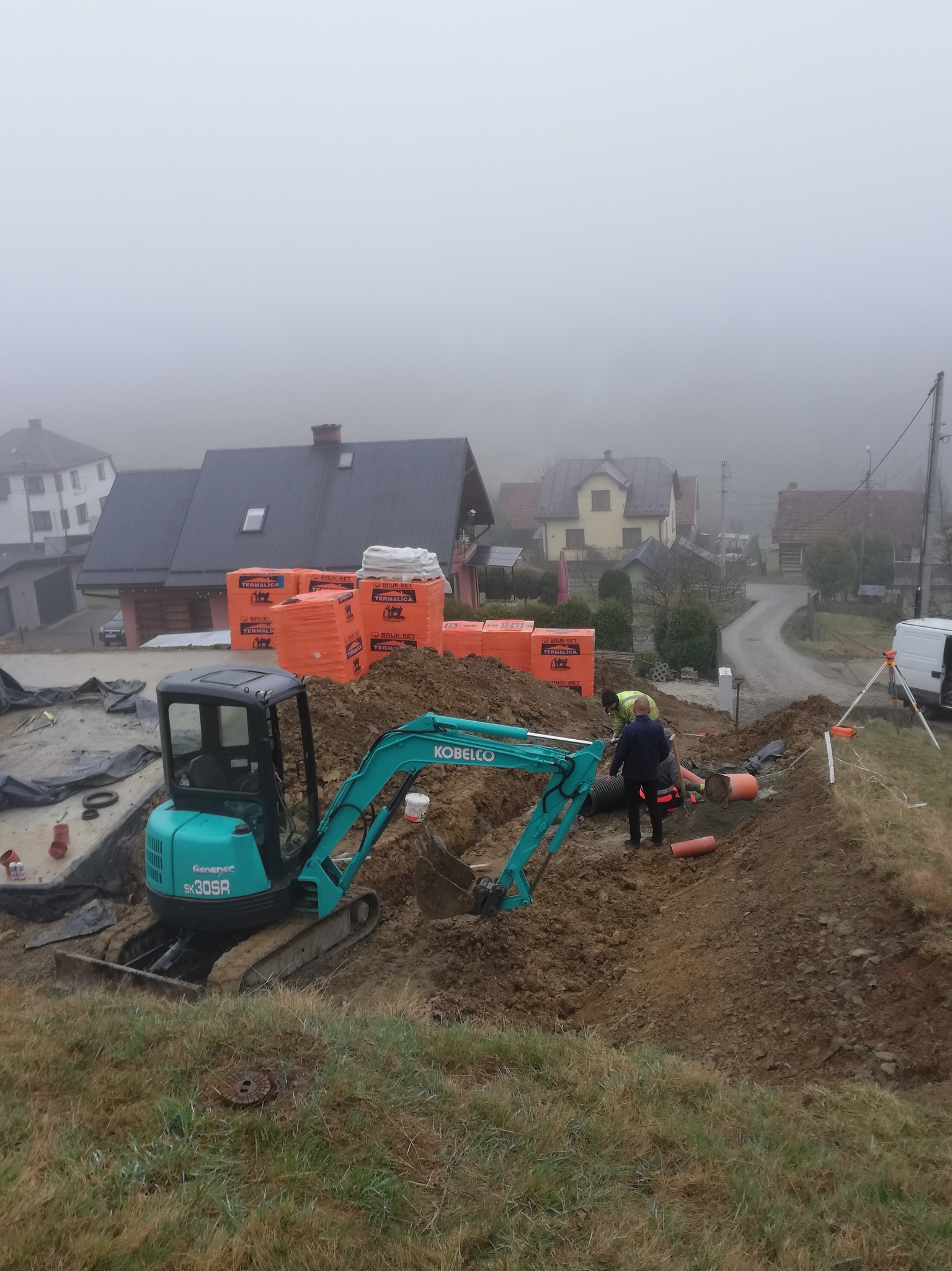 Niebieska minikoparka Kobelco SK30SR na placu budowy w mglisty dzień, obok sterty pomarańczowych bloczków TERMALICA, pracownicy układają rury w wykopie, w tle zabudowa jednorodzinna i sprzęt...