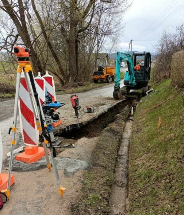 Wąski wykop wzdłuż drogi, mała koparka i wywrotka w tle, niwelator na trójnogu na pierwszym planie, narzędzia budowlane obok