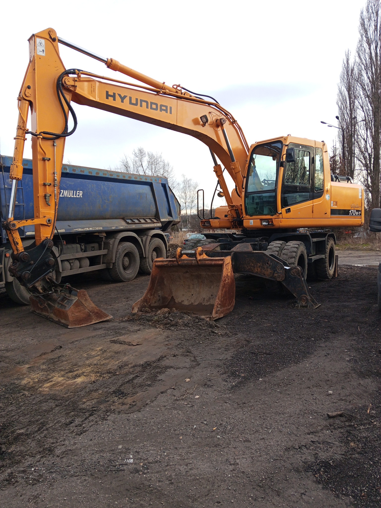 Żółta koparka kołowa Hyundai 170W-7 z łyżką na błotnistym terenie, obok niebieska ciężarówka Zmuller. Sprzęt budowlany w trakcie pracy.