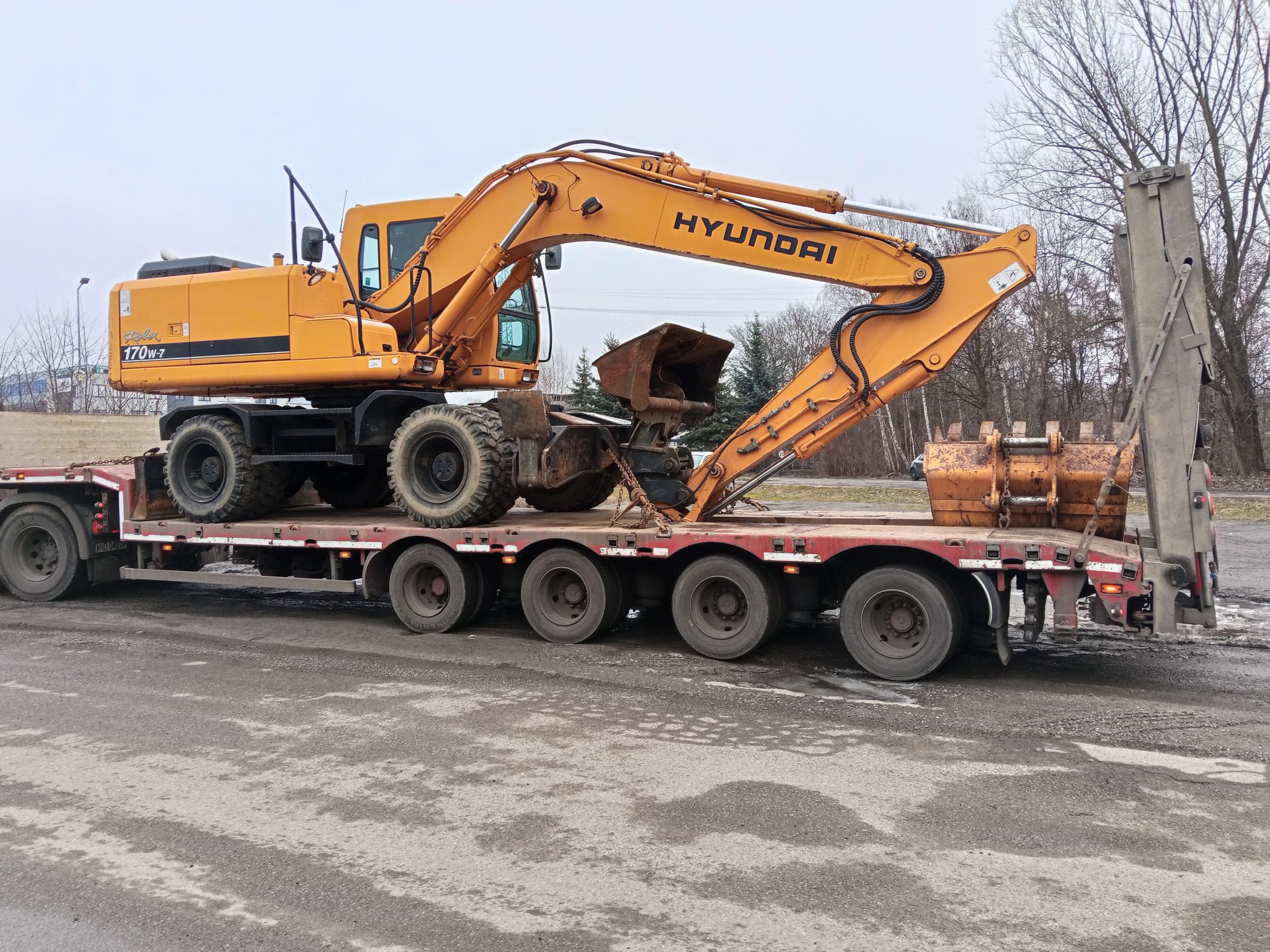 Żółta koparka kołowa Hyundai 170W-7 na niskopodwoziowej lawecie. Widoczne łyżki koparki. Transport maszyny budowlanej na plac budowy. Pochmurne niebo w tle.