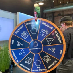 Mężczyzna w czapce pije drinka przy kole fortuny z napisem 'drinks2cash.com' i polami 'Duży Prezent', 'Mały Prezent', 'Drink', 'Spróbuj jeszcze raz', 'Nie tym razem'.