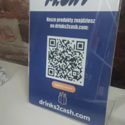 Stojak reklamowy z logo Float, adresem drinks2cash.com i kodem QR zachęcającym do skanowania i zamawiania, umieszczony na białym blacie na tle ceglanej ściany.