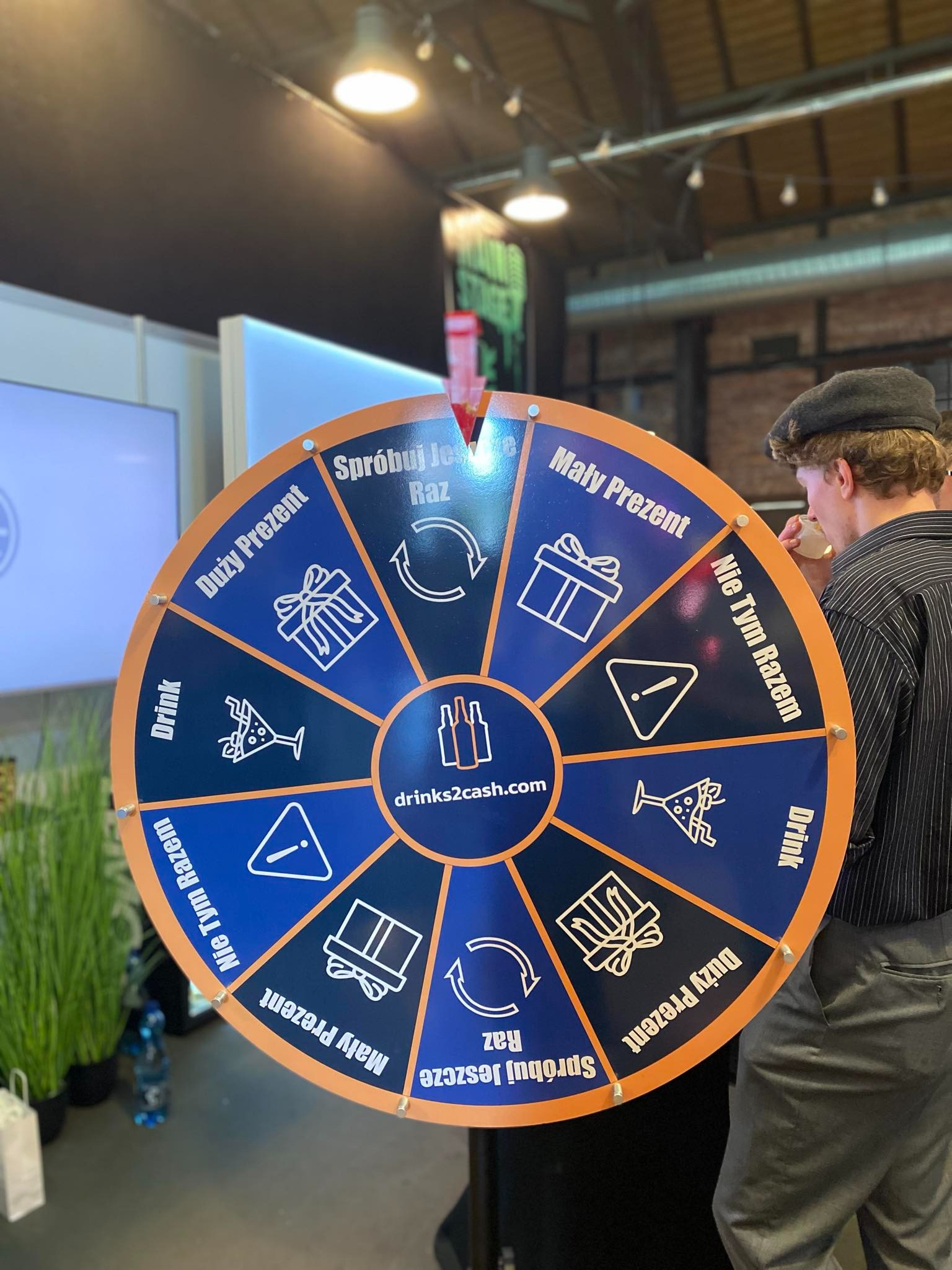 Mężczyzna w czapce pije drinka przy kole fortuny z napisem 'drinks2cash.com' i polami 'Duży Prezent', 'Mały Prezent', 'Drink', 'Spróbuj jeszcze raz', 'Nie tym razem'.