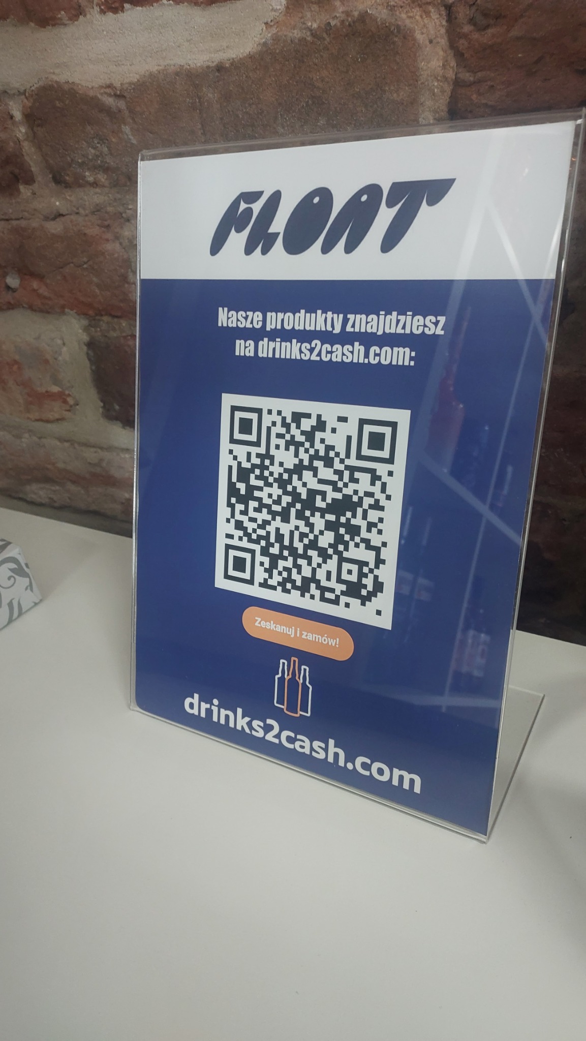Stojak reklamowy z logo Float, adresem drinks2cash.com i kodem QR zachęcającym do skanowania i zamawiania, umieszczony na białym blacie na tle ceglanej ściany.