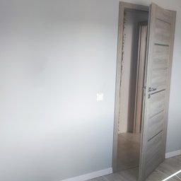 Doradztwo i obsługa techniczna Radosław Wojtczak - Świeżo pomalowane szare ściany w pokoju z jasną podłogą z paneli, biała listwa przypodłogowa i otwarte drewniane drzwi z szarą okleiną.