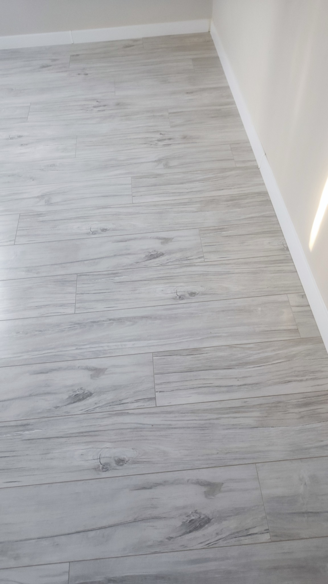 Jasnoszare panele podłogowe imitujące drewno, ułożone wzdłuż ściany z białą listwą przypodłogową, widoczne delikatne usłojenie i sęki na powierzchni paneli.