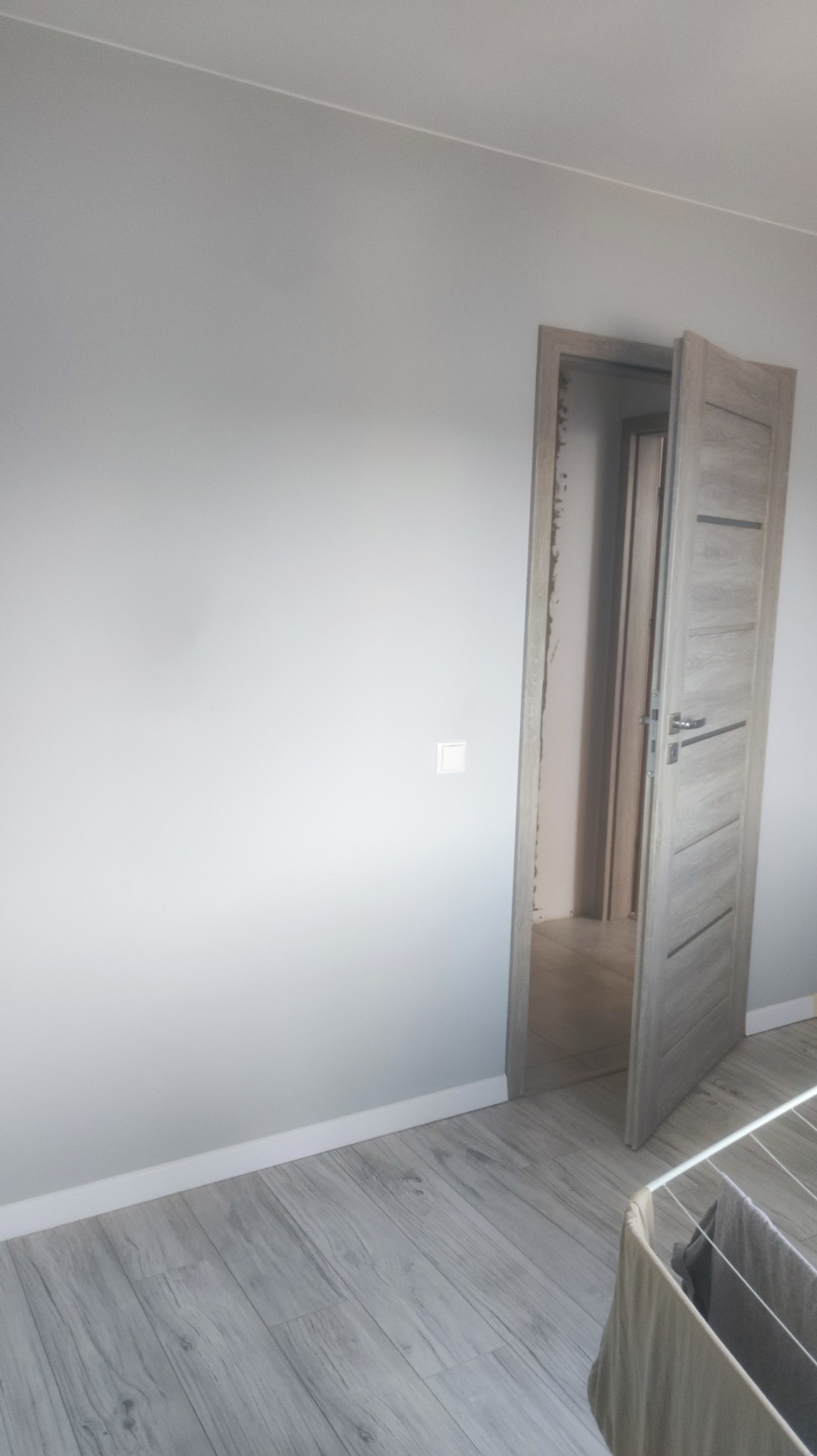 Świeżo pomalowane szare ściany w pokoju z jasną podłogą z paneli, biała listwa przypodłogowa i otwarte drewniane drzwi z szarą okleiną.