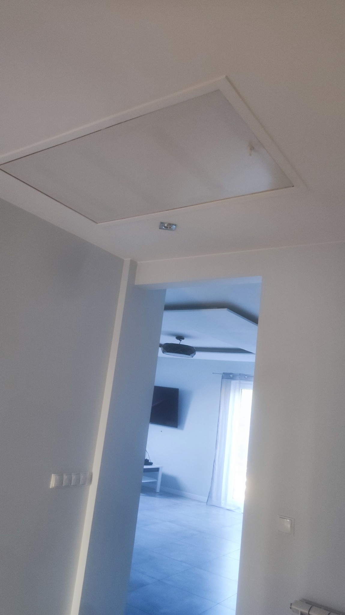 Białe, świeżo pomalowane wnętrze mieszkania z widocznym włazem na suficie i fragmentem salonu w tle.