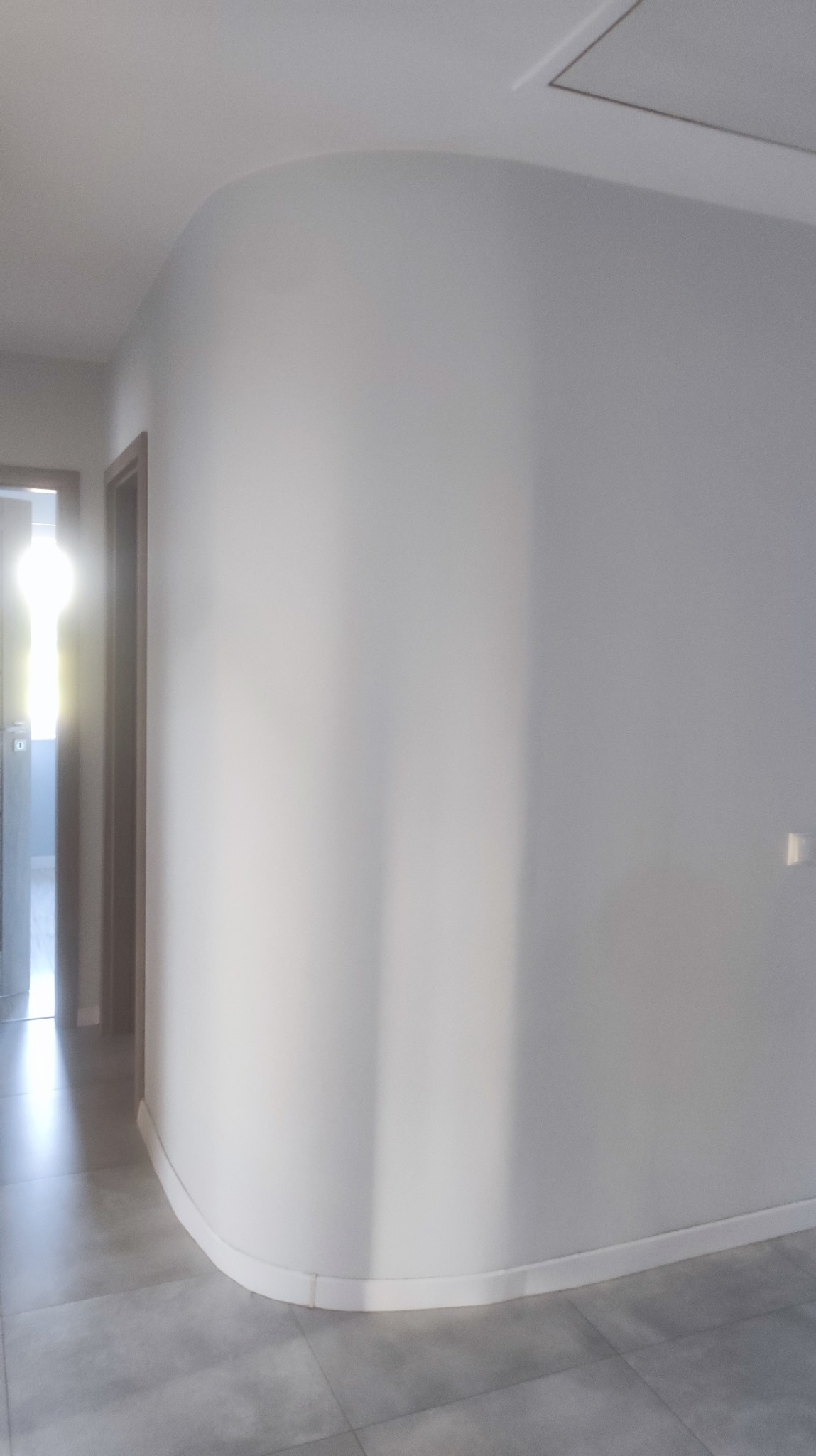 Świeżo pomalowane, jasne wnętrze w Warszawie z zaokrągloną ścianą i szarą podłogą z płytek, widok na drzwi z ościeżnicami. Ściana oświetlona padającym światłem.