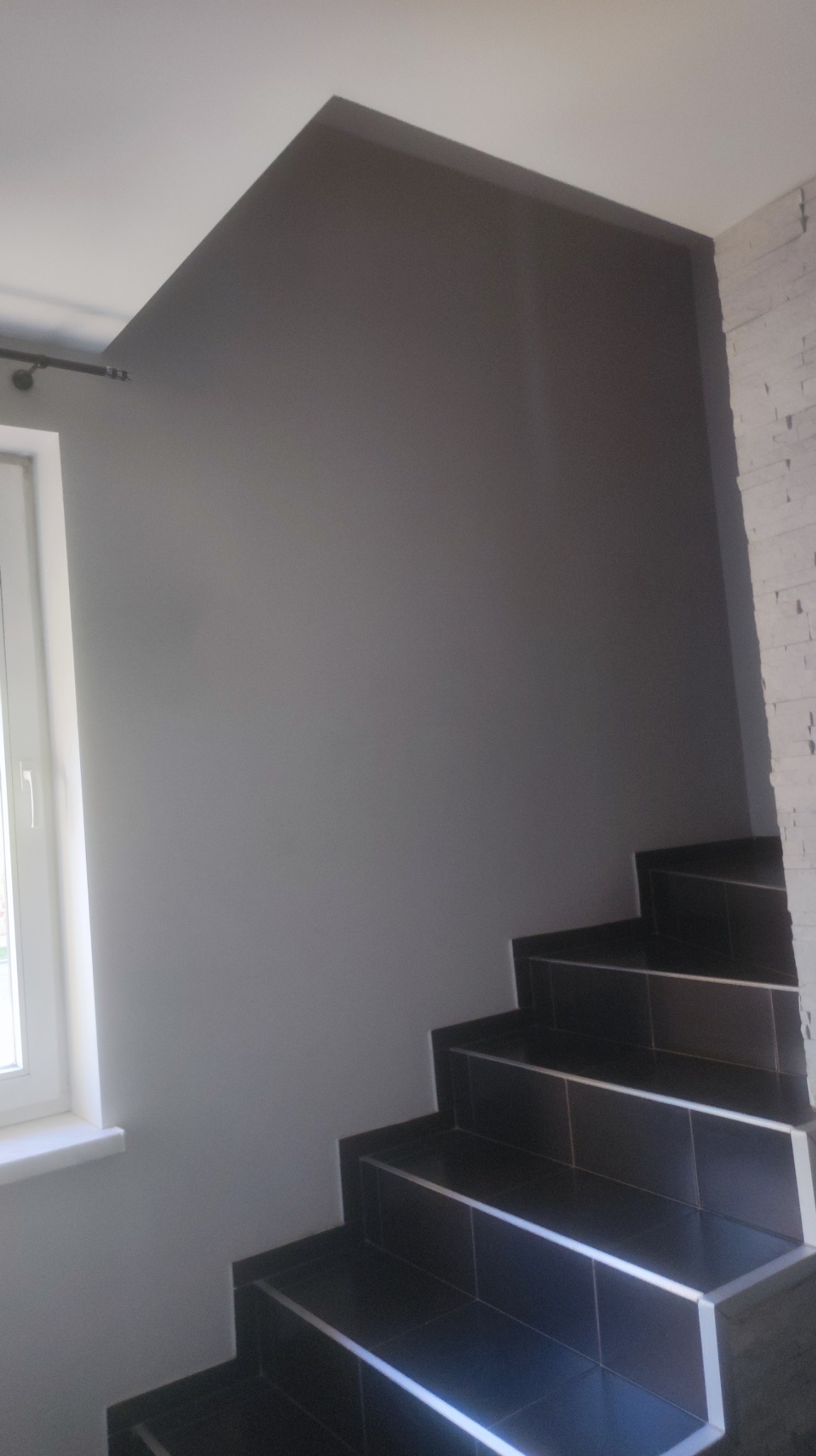 Klatka schodowa po remoncie: ściana pomalowana na szaro, schody wyłożone ciemnymi płytkami z metalowymi listwami, widoczne okno z białym parapetem i fragment ściany wyłożonej białą cegłą.