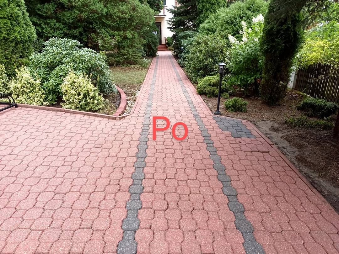 Czerwona kostka brukowa ułożona w ścieżkę z szarymi akcentami, prowadząca przez zadbany ogród z różnorodną roślinnością i lampą ogrodową.