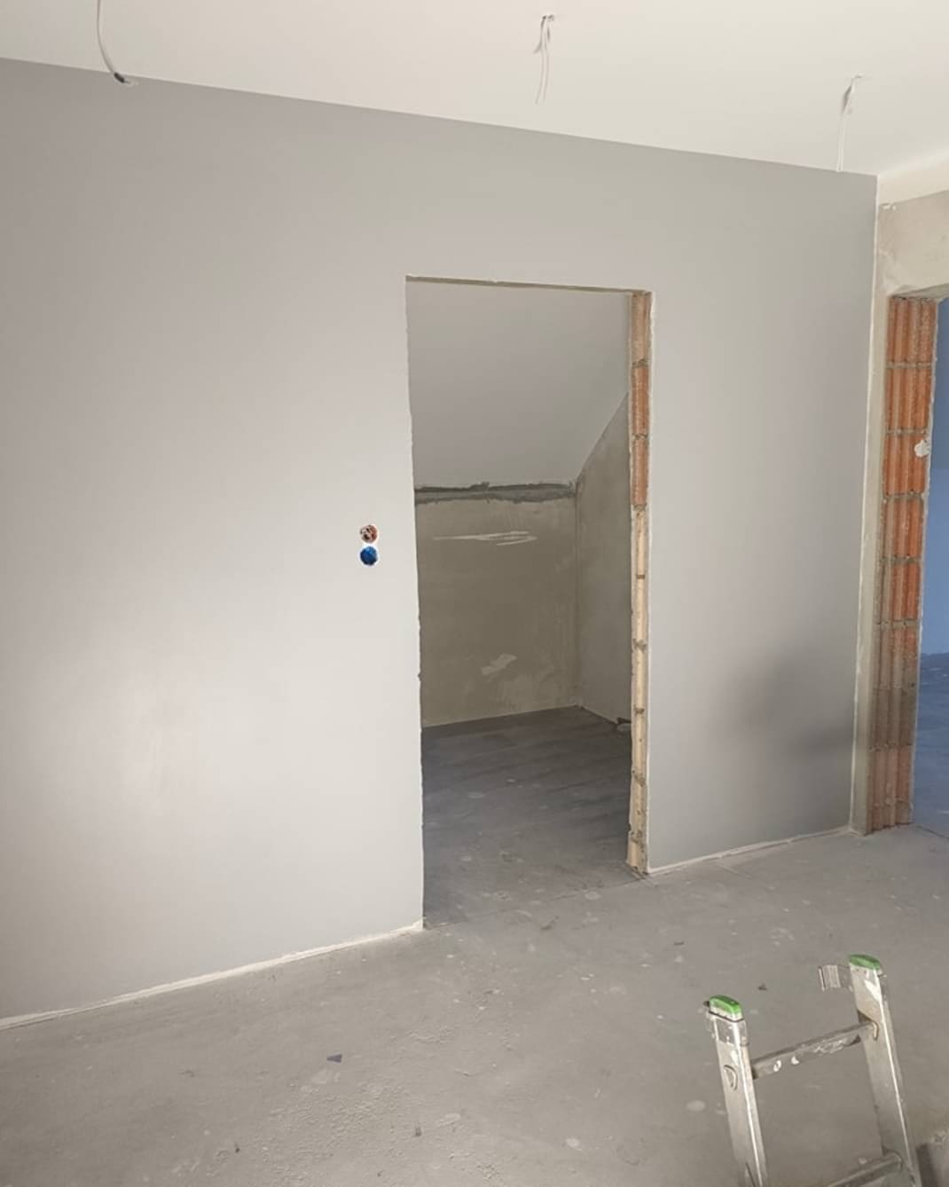 Wnętrze w trakcie remontu: świeżo pomalowane szare ściany, widoczne instalacje elektryczne, surowy beton na podłodze, aluminiowa drabina oparta o ścianę, otwór drzwiowy bez ościeżnicy.