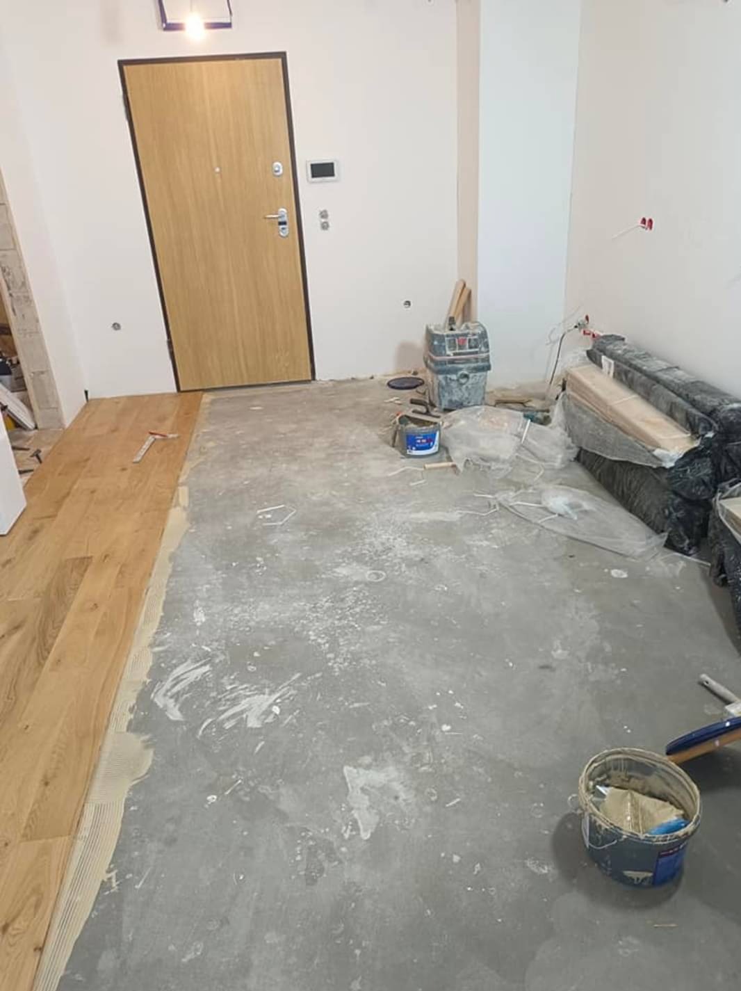 Fragment pokoju w trakcie układania jasnych paneli podłogowych, widoczny klej, paczki paneli w folii, narzędzia i wiadro na podłodze.